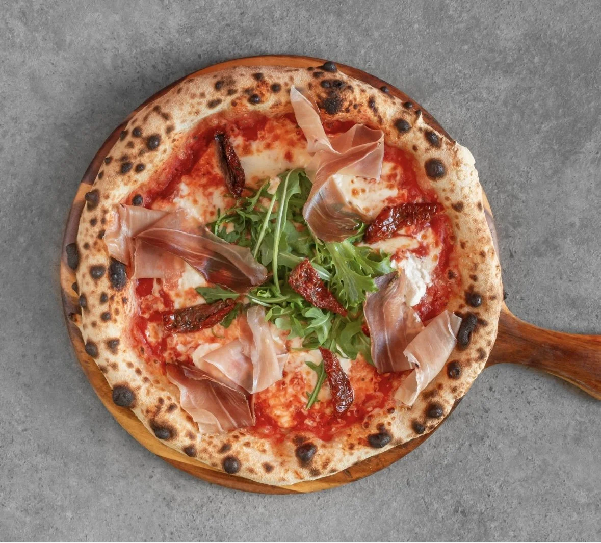 Neapolitan pizza with melted mozzarella cheese, sun-dried tomatoes, fresh arugula, and prosciutto, on a wooden pizza board.