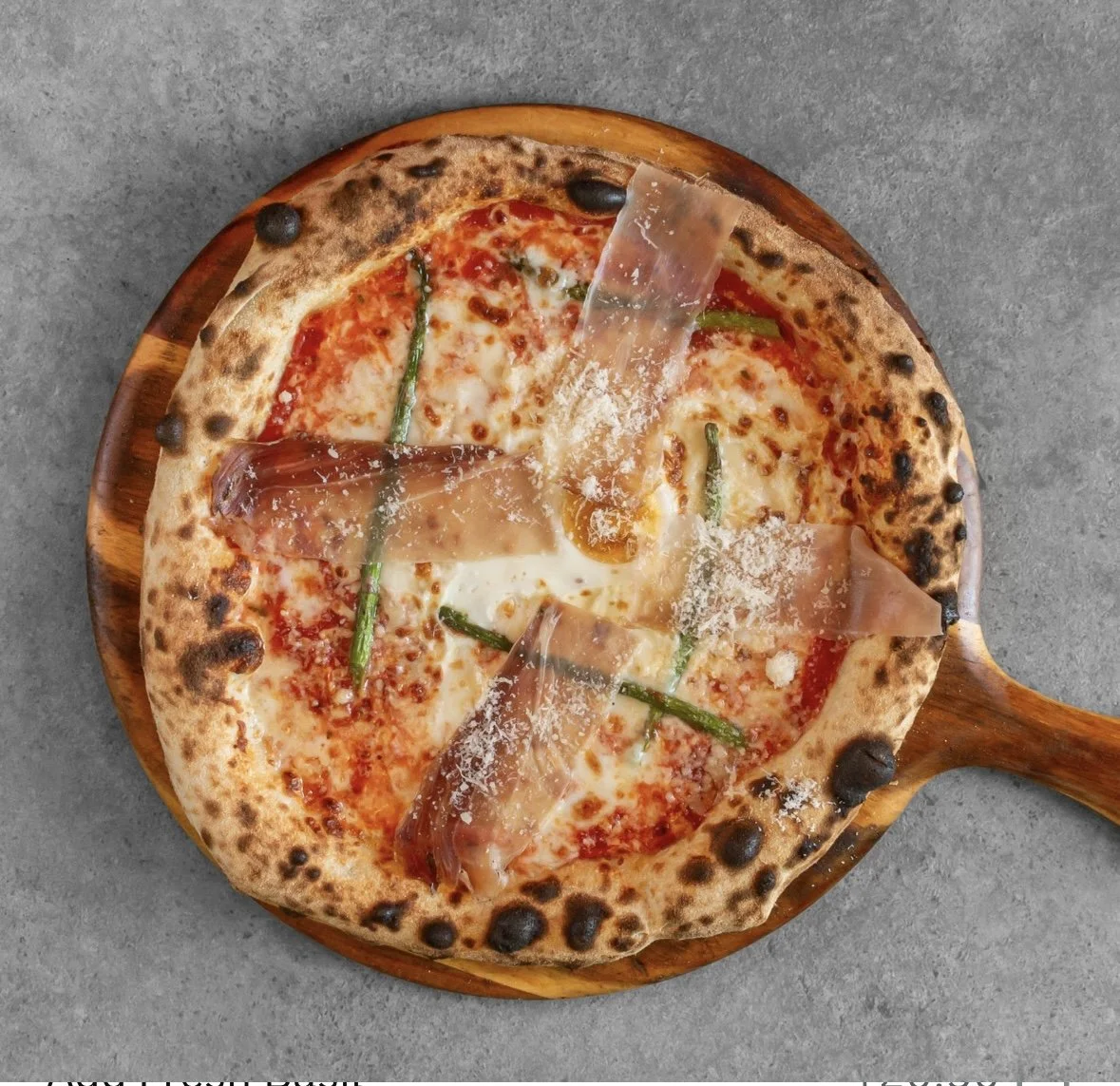 Pizza with cheese, green peppers, and slices of prosciutto on a wooden paddle.