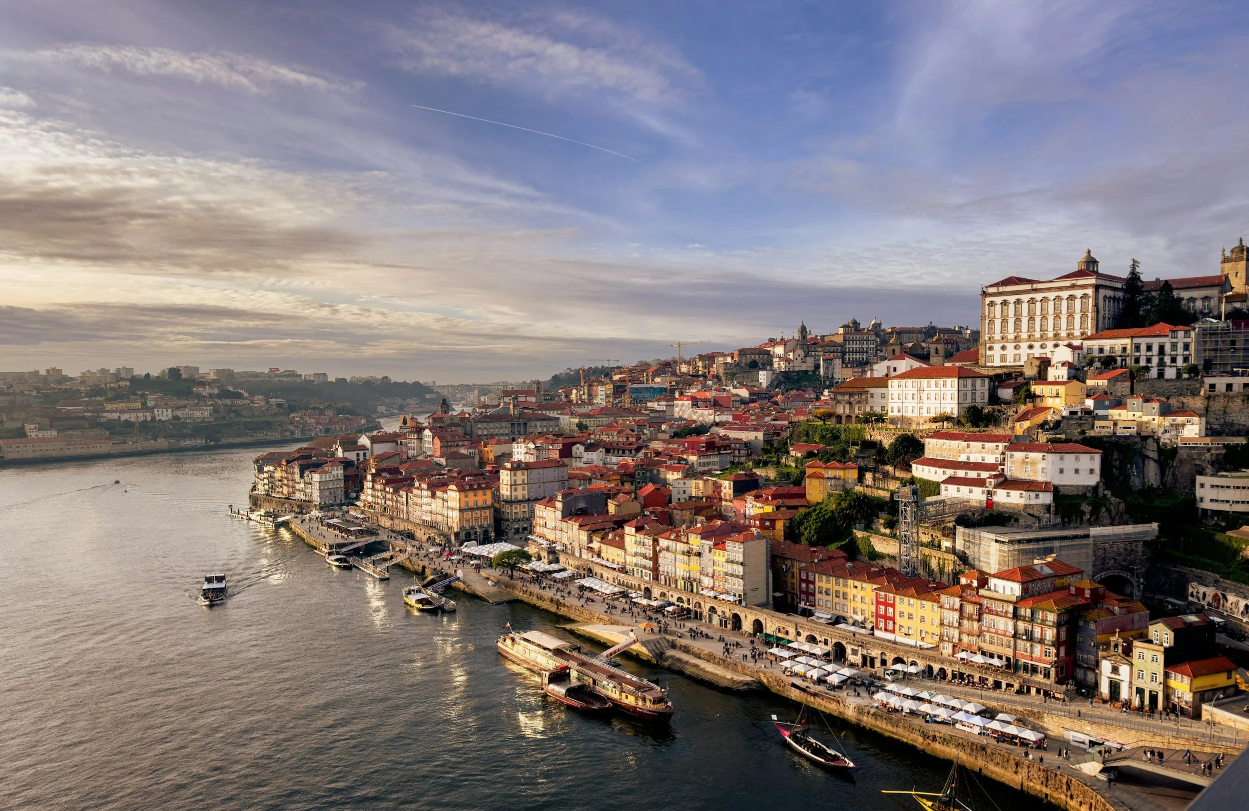 View of a colorful riverside town with boats on the river, hillside buildings, and historic architecture under a partly cloudy sky.