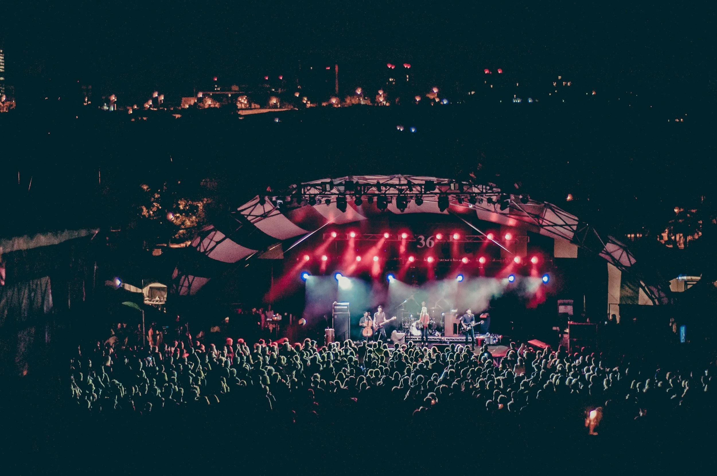An outdoor concert at night with a large crowd watching a band perform on stage under colorful lights.