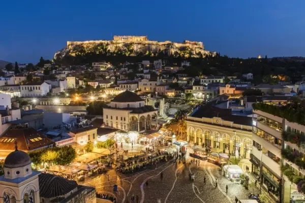 Nighttime view of a historic city with a castle on a hilltop, illuminated buildings, and a lively outdoor market or square with people walking and lights.