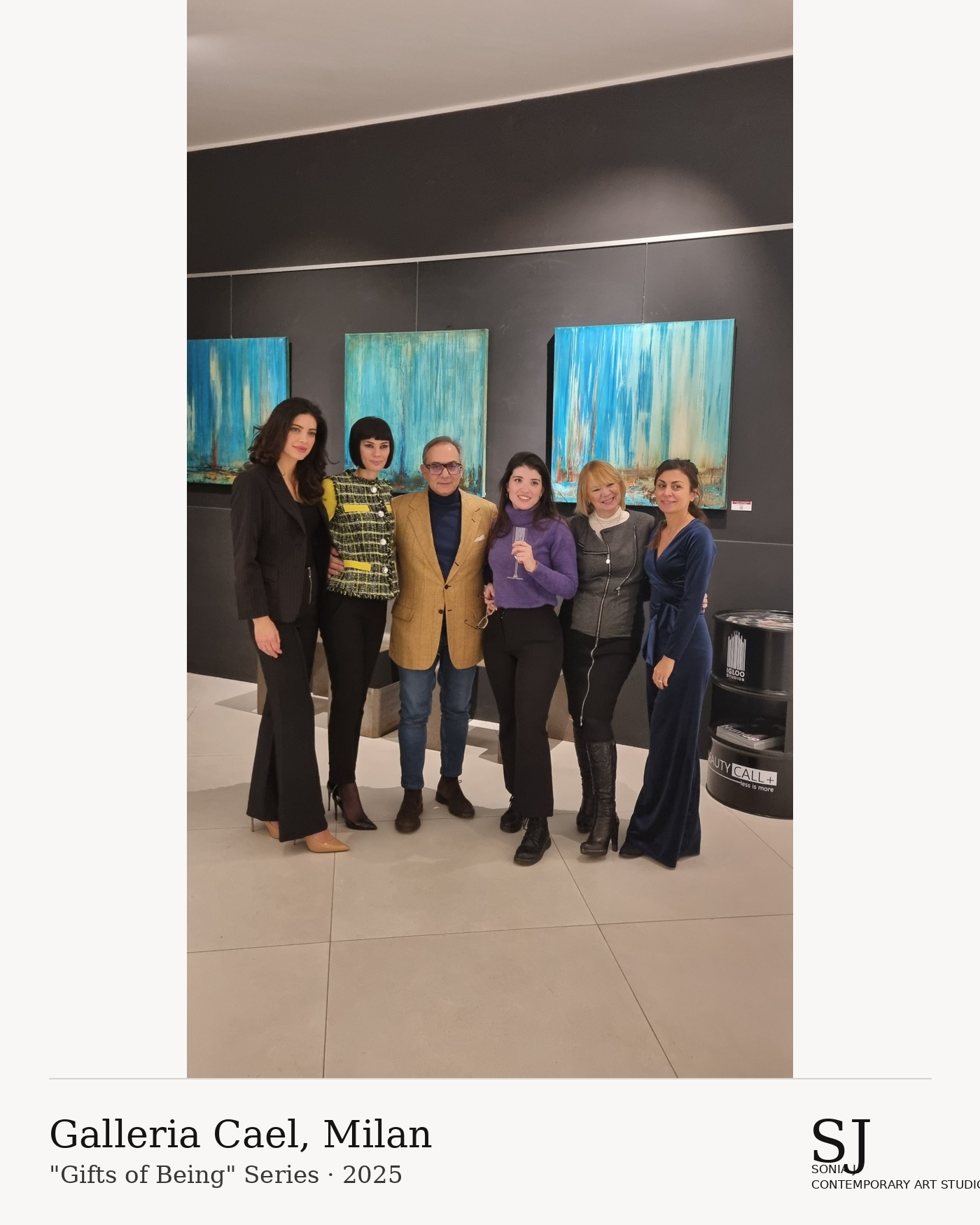 Six people standing together in an art gallery, posing for a photo. Behind them are abstract paintings with blue-green tones on a dark gray wall. There is a black stand with promotional materials in the lower right corner. The gallery is well-lit, an