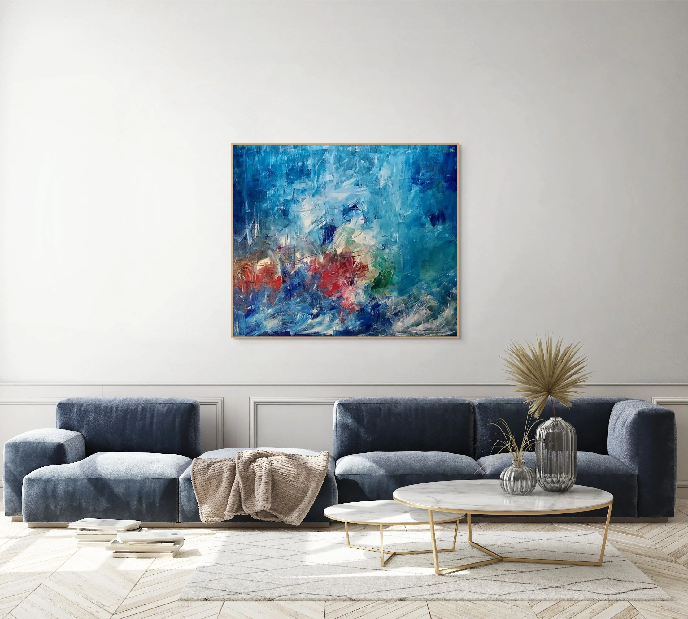 Living room with a large abstract painting in shades of blue and red on the wall above a dark blue sofa. The sofa has a beige blanket draped over one arm. Two marble-topped coffee tables with gold legs are in front, decorated with glass vases and dried plants. The room features off-white walls and light wood flooring, with a subtle geometric pattern rug.