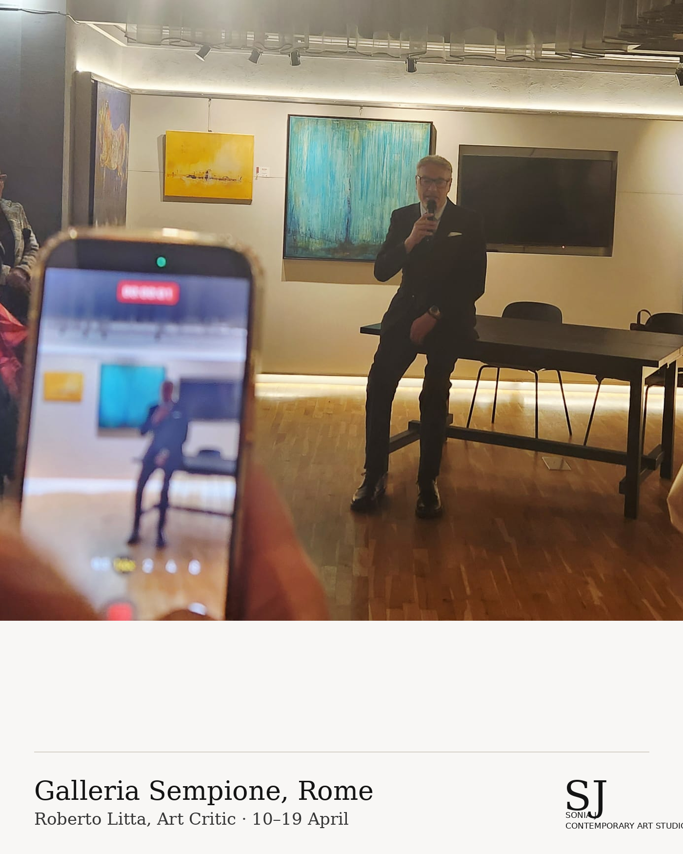 A man in a suit is sitting on a table, holding a microphone in a gallery, with colorful abstract paintings on the wall behind him. A large television is mounted on the wall to his right, and a person in the foreground is taking a photo of him with a 