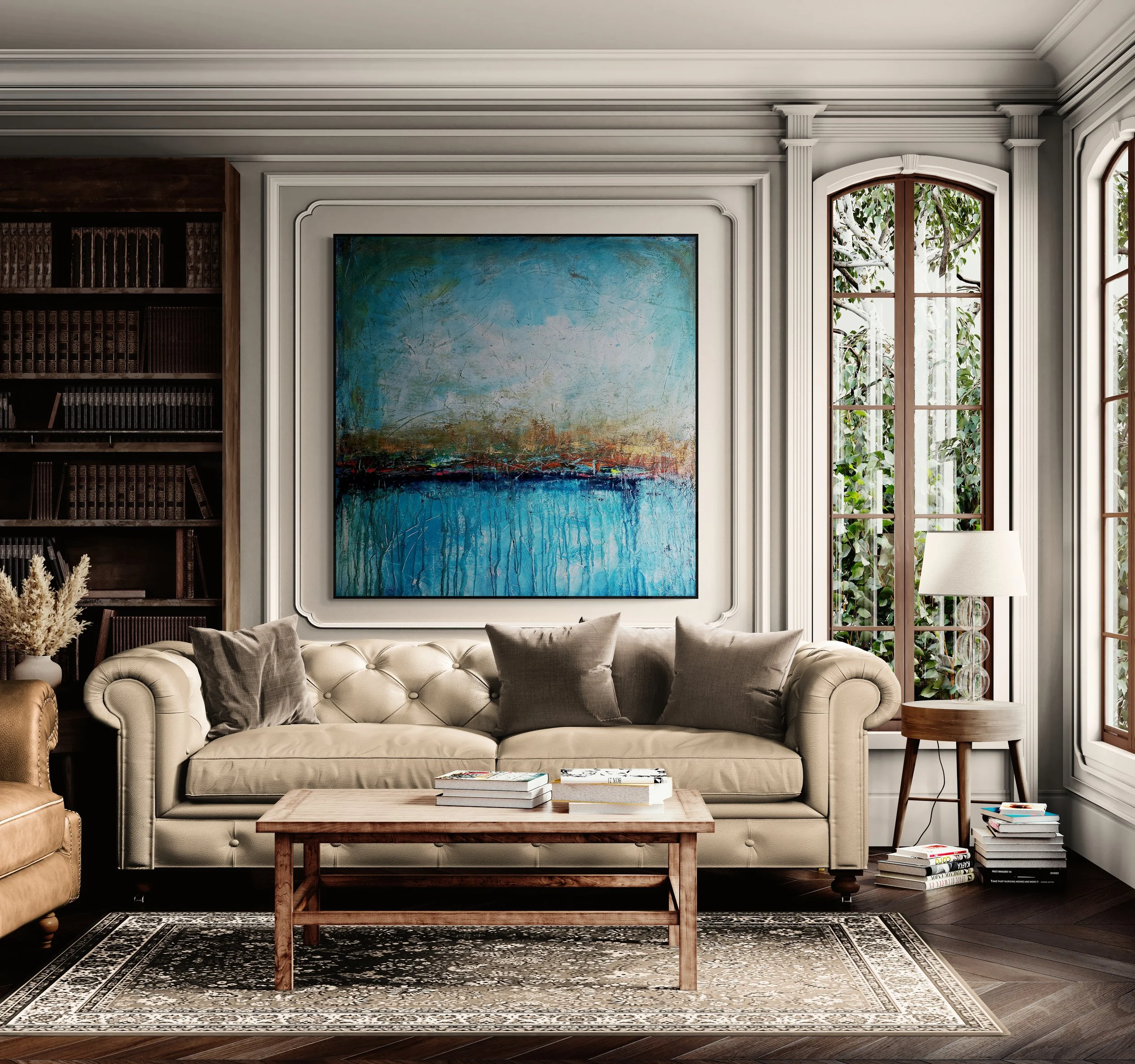 Living room with a beige tufted sofa, gray pillows, a wooden coffee table with books, a patterned rug, a wooden side table with a lamp, a large abstract painting, bookshelf, and tall windows with greenery outside.