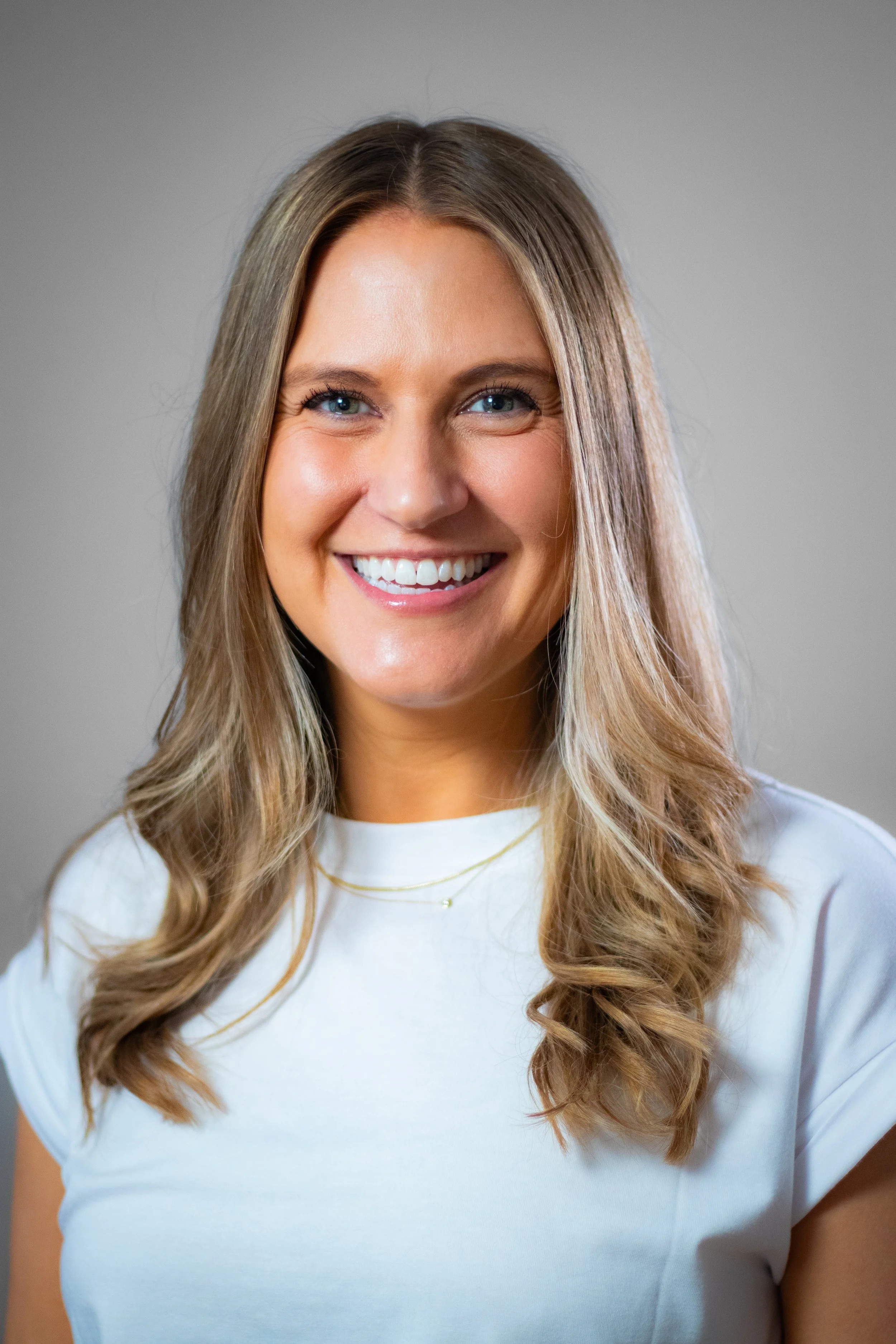 Portrait of a smiling woman with long, wavy blonde hair, wearing a white top, and standing against a neutral background.