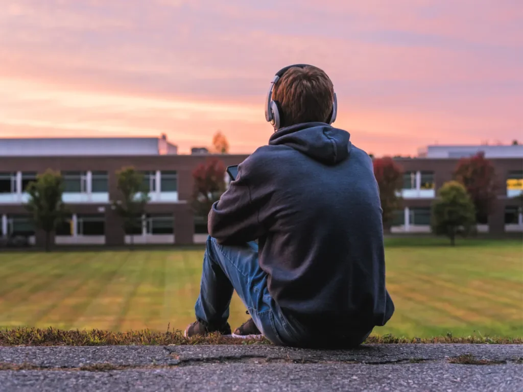 A person sitting on a curb with headphones, looking at a sunset over a grassy area and modern building in the background.