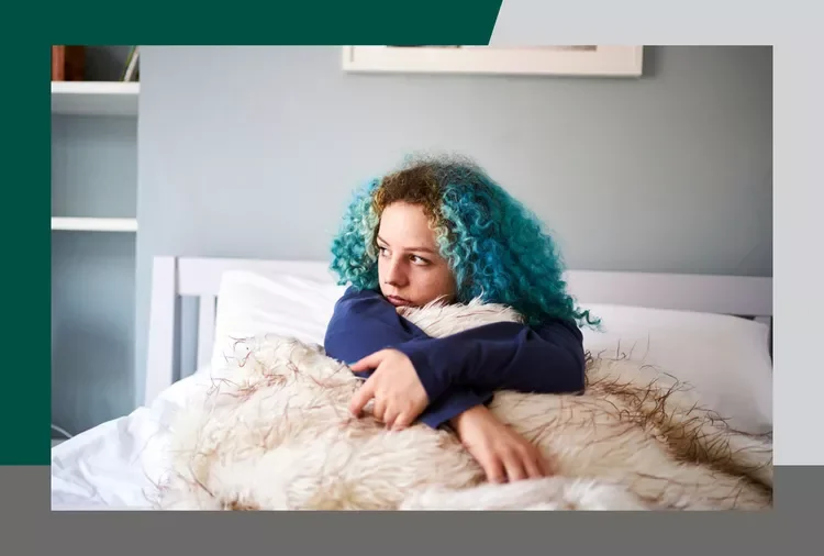 Young woman with blue curly hair sitting on bed, hugging a fluffy beige pet.
