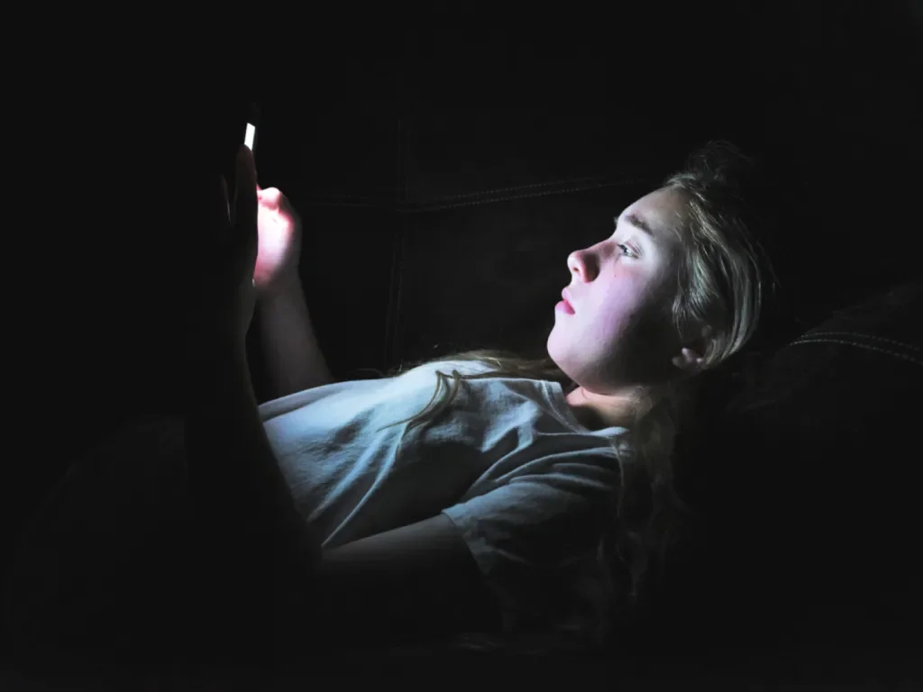 A young girl lying in bed in darkness, illuminated by the light of a smartphone she is holding in her hands.