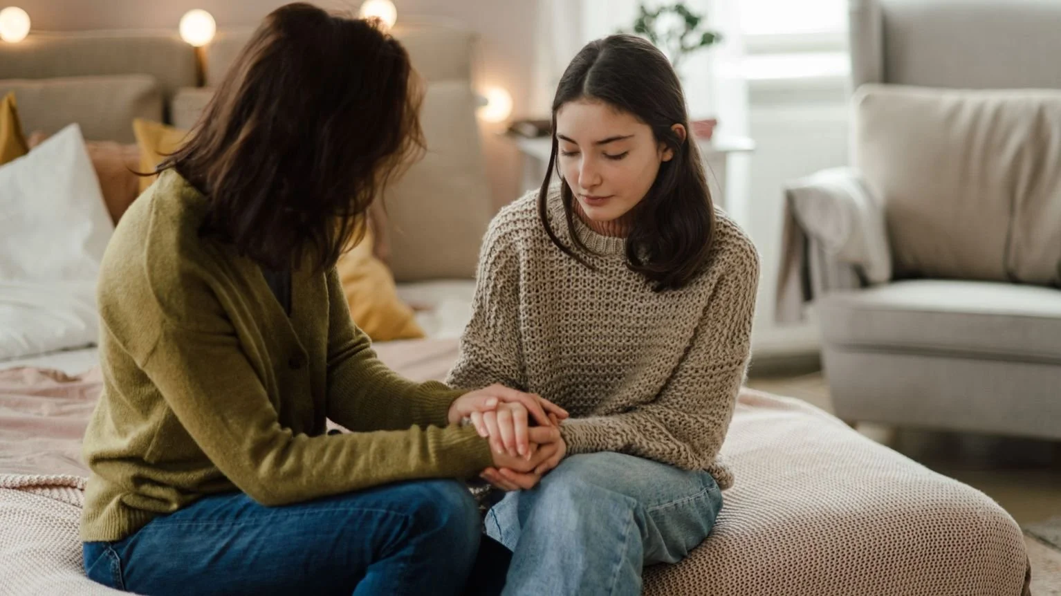 Two women sit close on a bed in a cozy, well-lit room, holding hands and sharing an emotional moment.