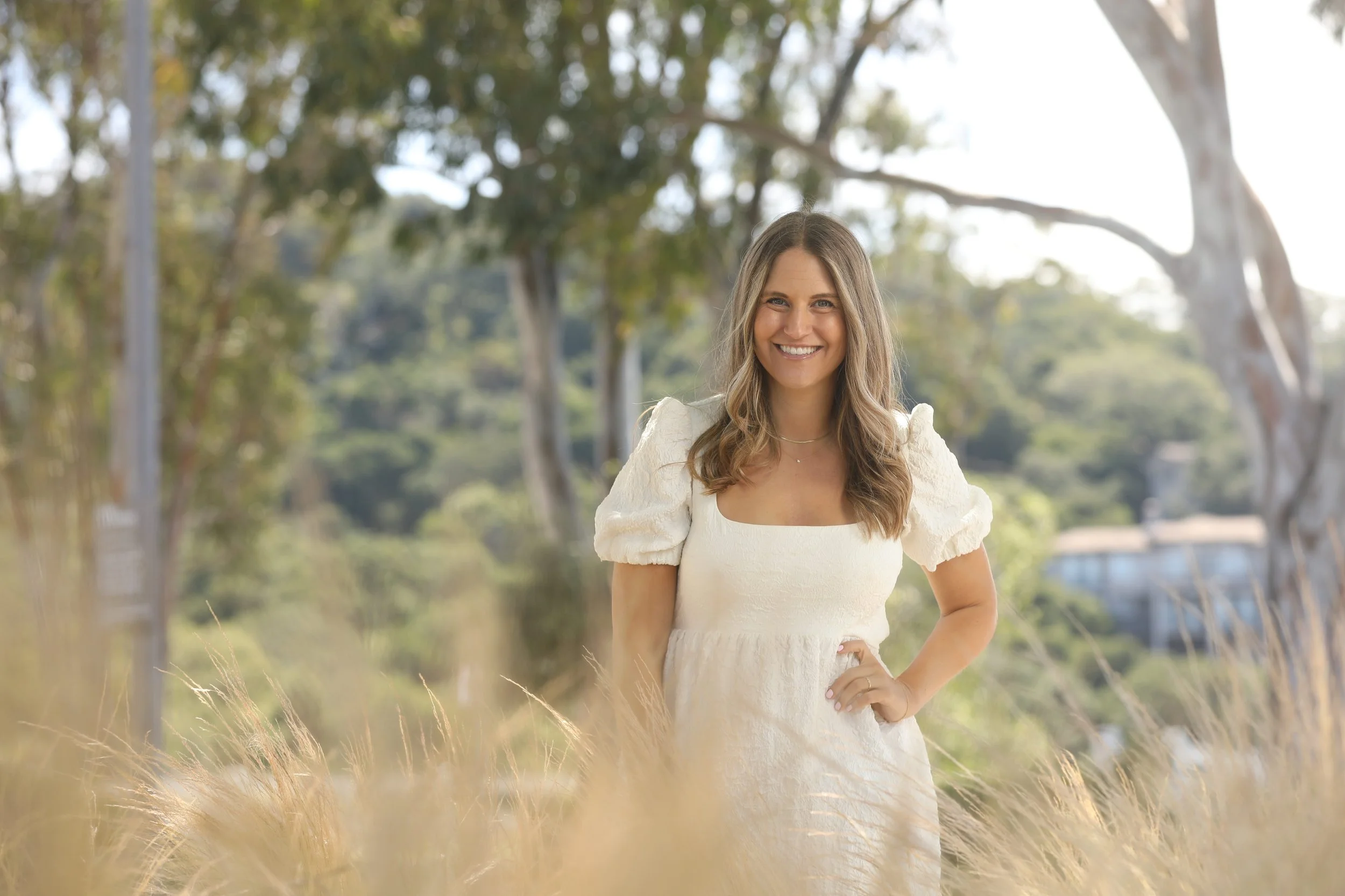 A smiling woman with long brown hair wearing a white dress with puffed sleeves standing outdoors among trees and tall grass.