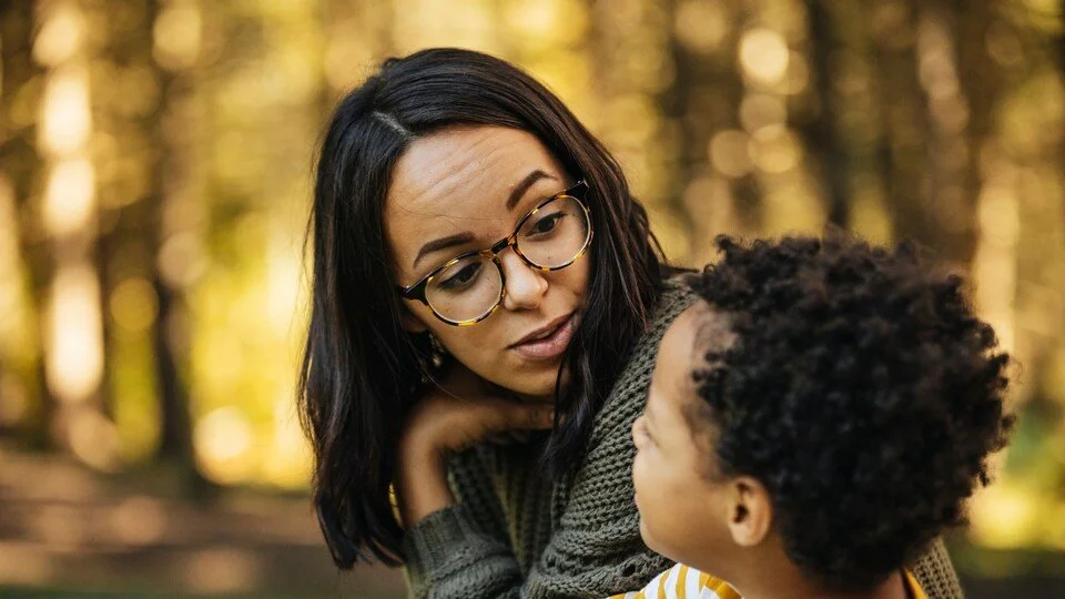 A woman with glasses and dark hair looks at a young child with curly hair, outdoors in a wooded area during daytime.