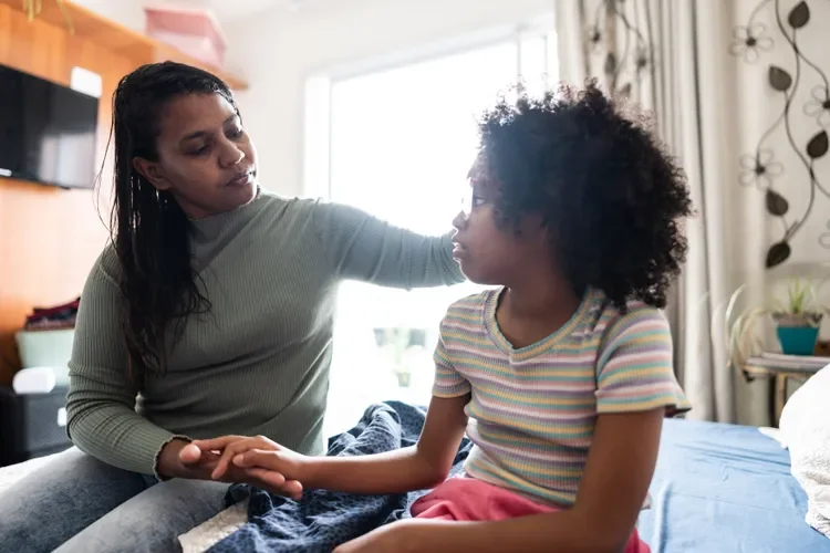 A woman comforting a young girl sitting on a bed in a bedroom with sunlight streaming in.