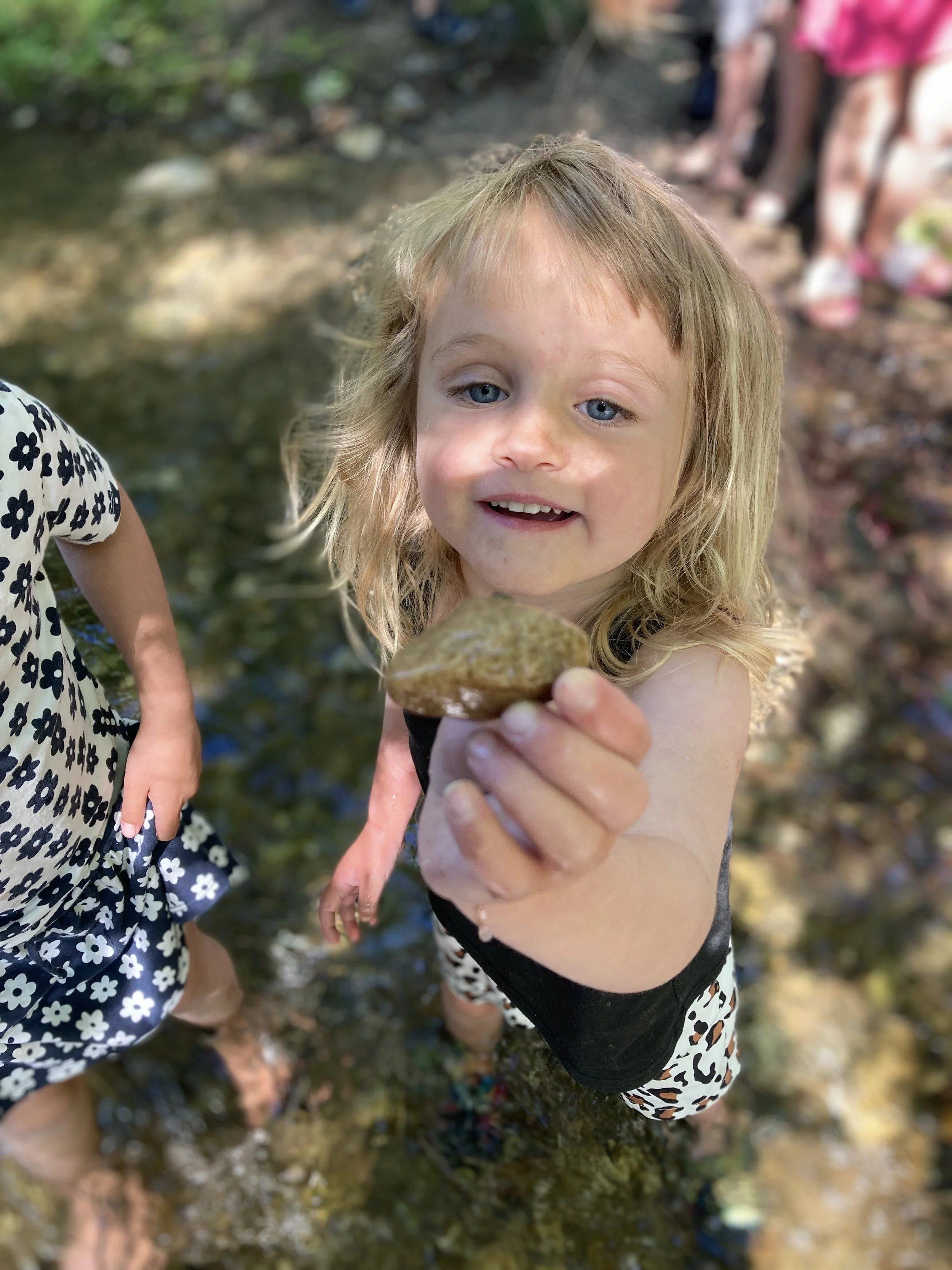 A young girl with blonde hair and blue eyes holding up a smooth stone while standing in a shallow creek with other children along the banks, in a wooded area.