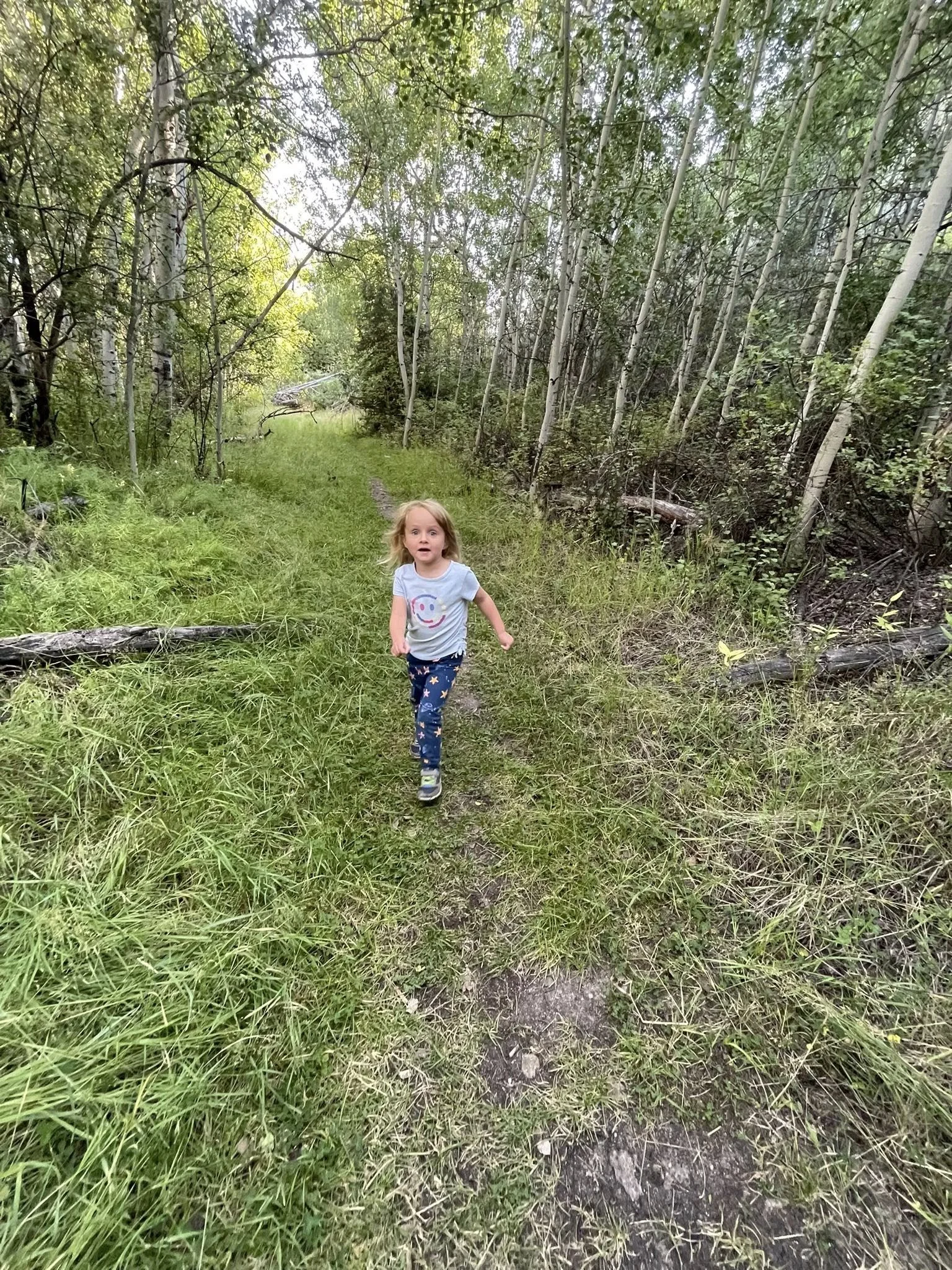 A young girl running on a narrow dirt trail through a green forest with tall trees and dense foliage.