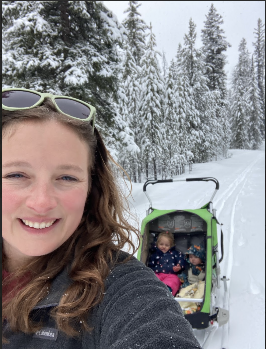 Smiling woman with sunglasses taking a selfie in a snowy forest, pulling a sled with two young children dressed in winter clothes.