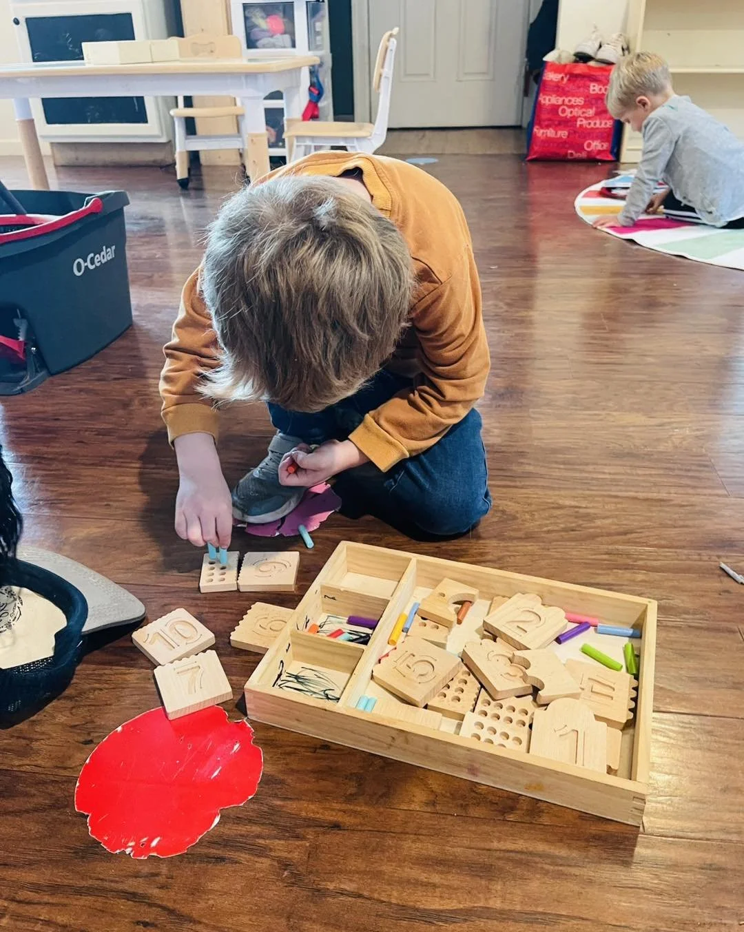 A young child kneeling on a wooden floor, playing with wooden number blocks and colorful markers in a playroom. Another child is in the background on a striped rug, also engaged in play.