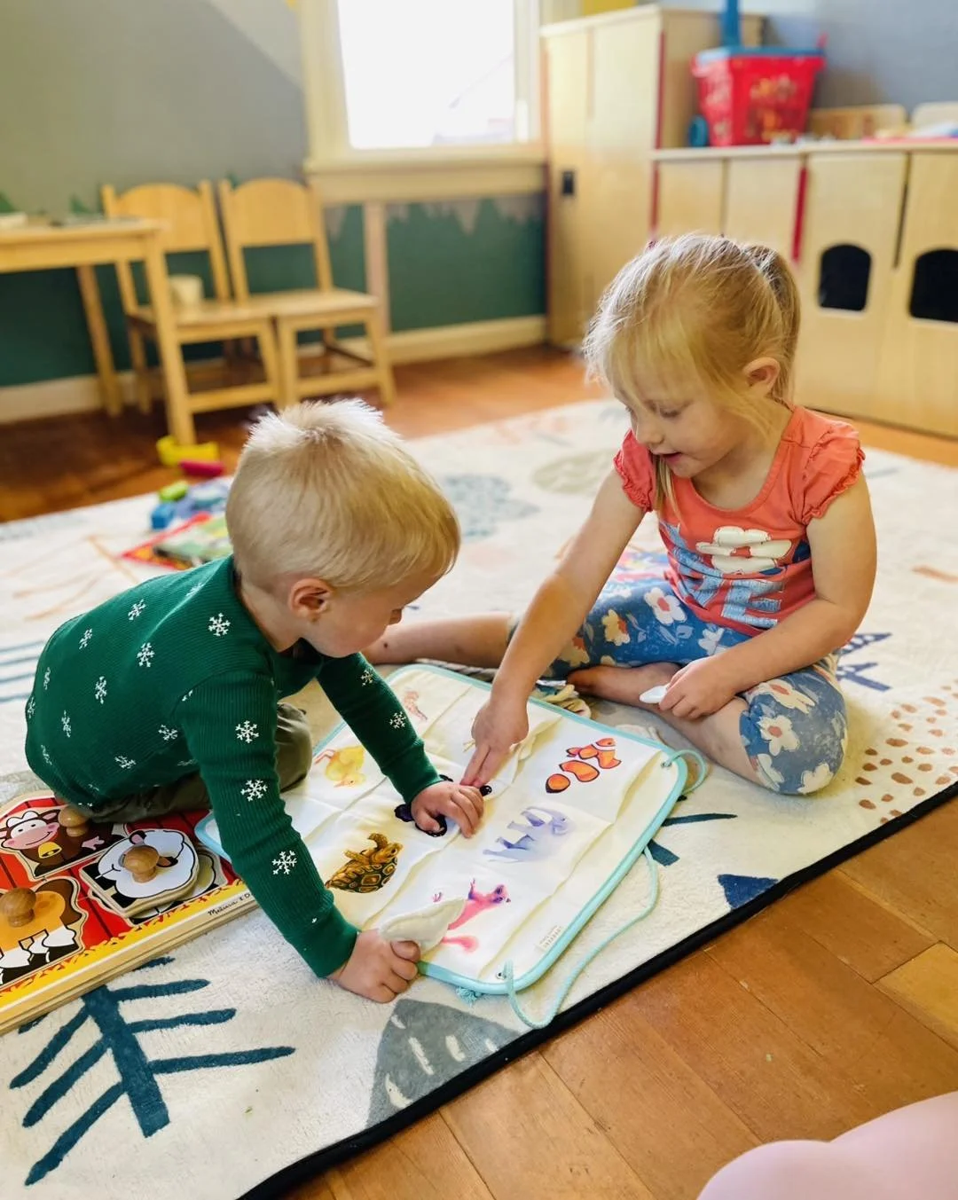 Two young children, a boy and a girl, sitting on a colorful play mat on a wooden floor, engaging with a soft activity book featuring animals and bright illustrations, in a playroom with wooden chairs and storage shelves in the background.