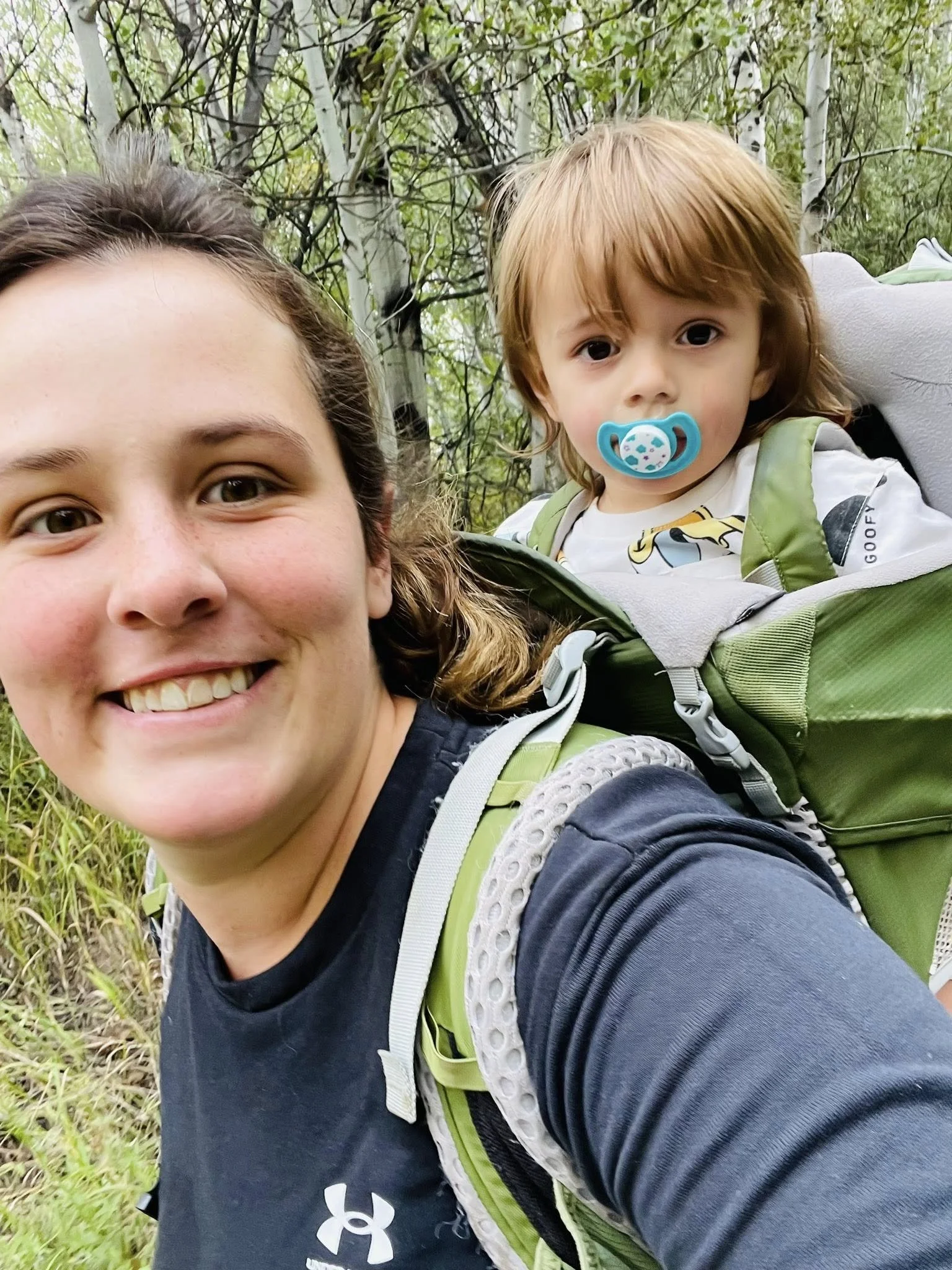 A woman with a backpack smiling while carrying a young child in a backpack carrier among trees in a forest.