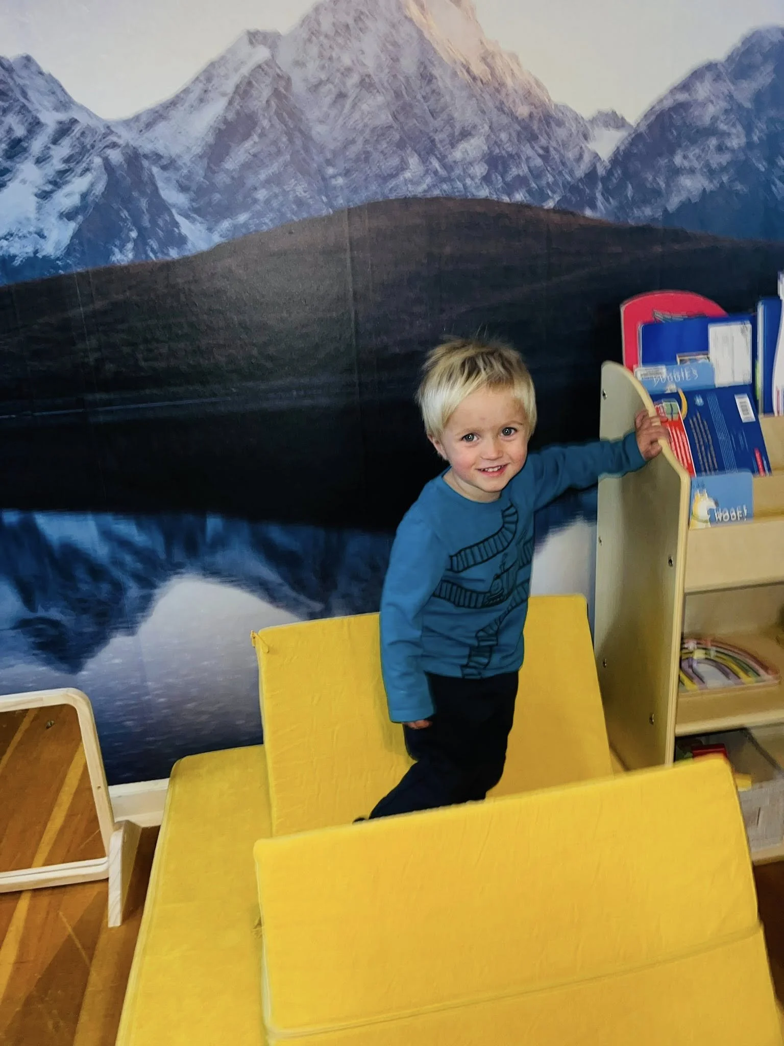 A young boy with blond hair wearing a blue long-sleeve shirt and dark pants standing inside a yellow cardboard box or playhouse, smiling at the camera. The background features a mural of a mountain landscape with snow-capped peaks and a reflective lake.