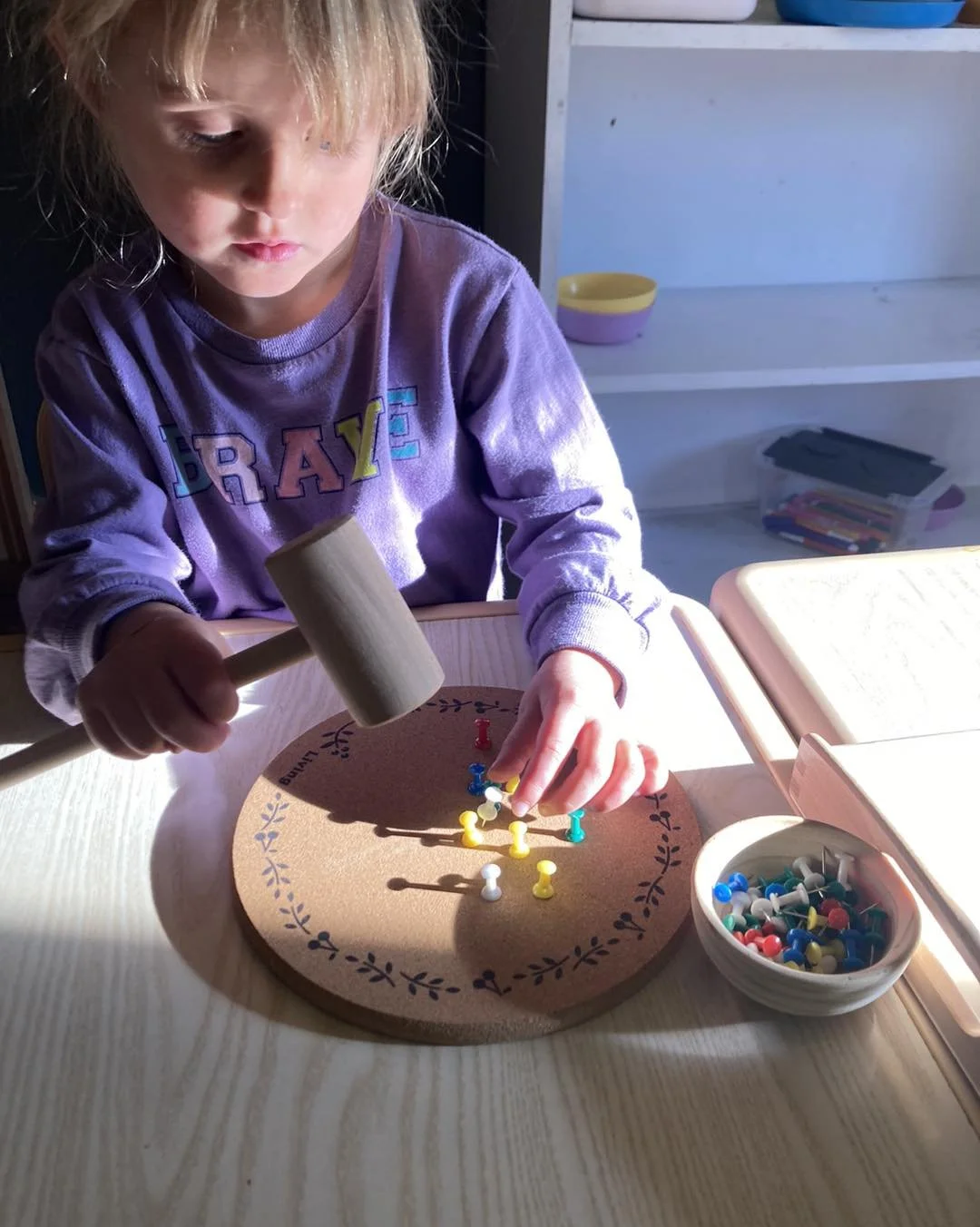 A young girl in a purple shirt with the word 'PRAISE' printed on it, using a small wooden hammer to place colorful pushpins on a round cork board with a decorative border.