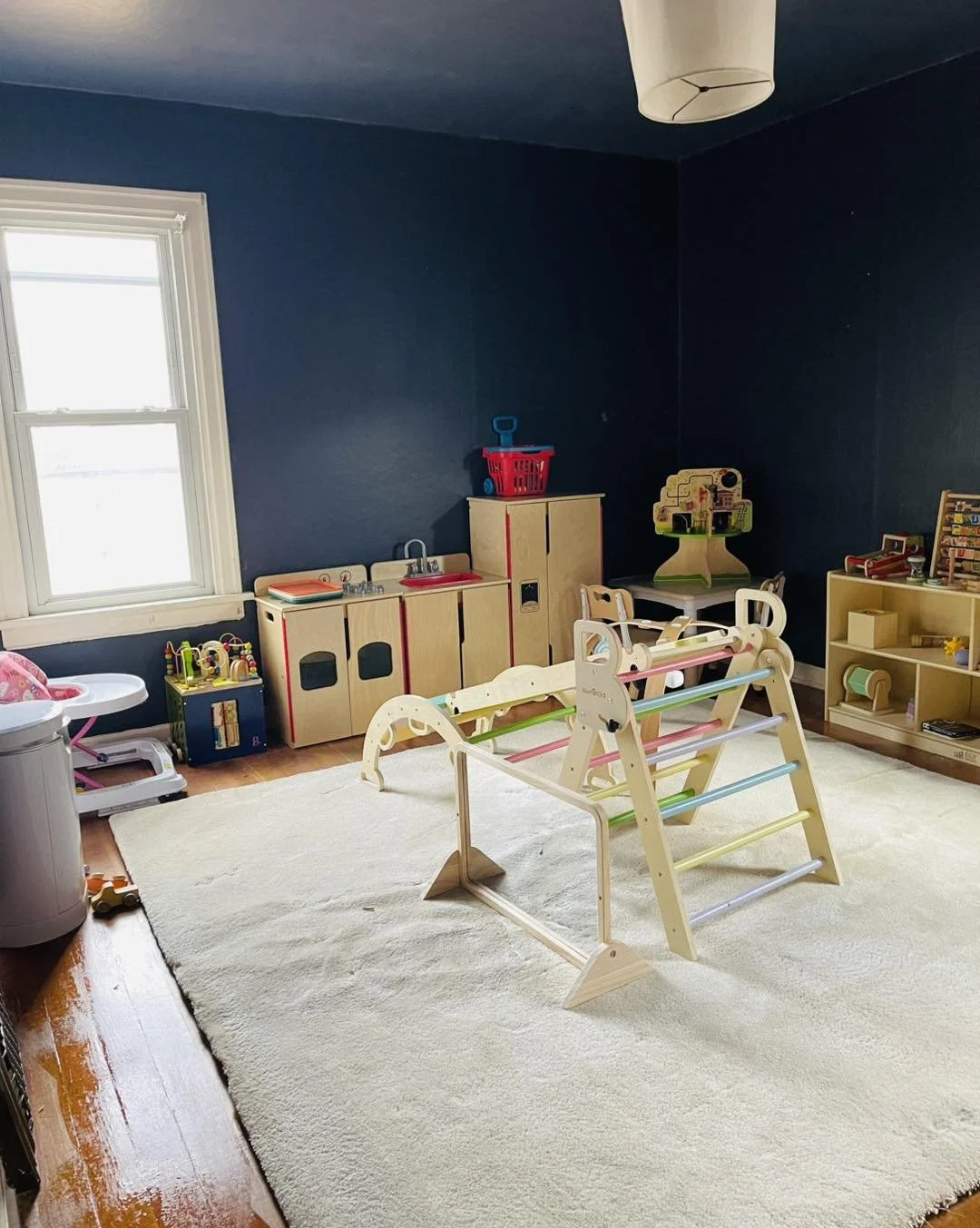 A children's playroom with dark blue walls, a large cream rug, a small play kitchen, a wooden climbing structure with colorful rungs, and various toys and shelves for storage.