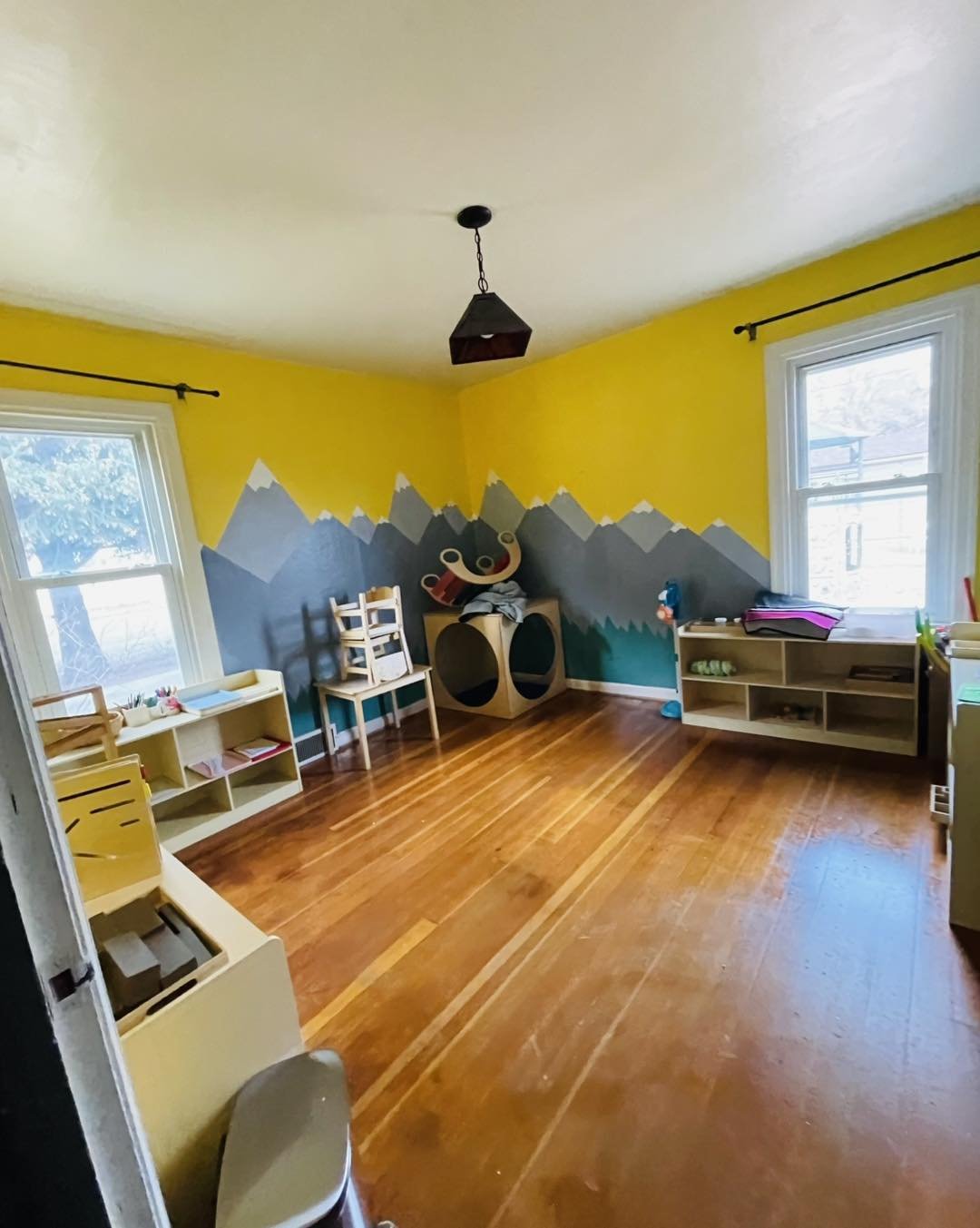 A children's playroom with yellow walls decorated with mountain mural, hardwood floor, and two large windows. There are shelves with toys and books, chairs, and a connected swan-shaped rocker.