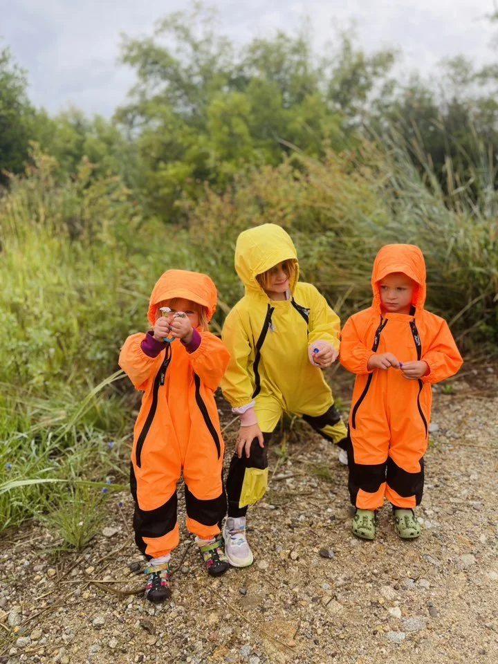 Three children in bright yellow and orange rain jackets and rain boots standing outdoors on a dirt path surrounded by greenery.