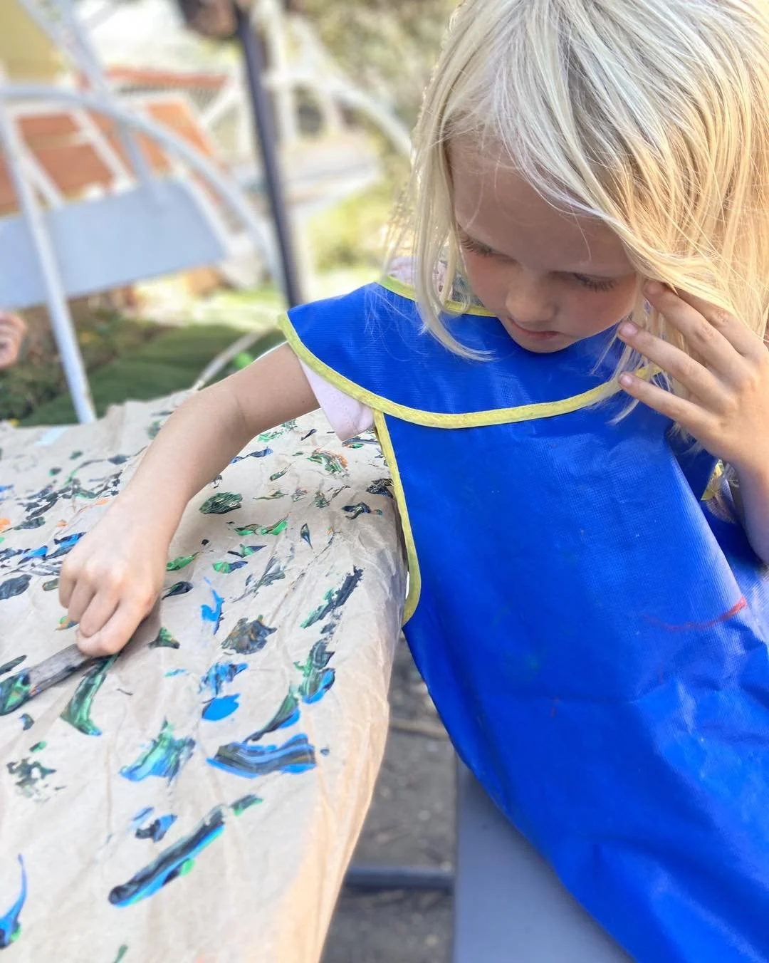 Young girl with blonde hair in a blue smock painted on a paper-covered table outdoors.