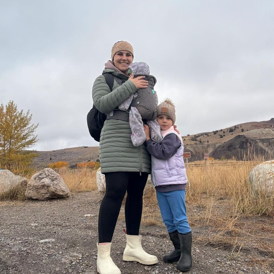 A smiling woman holding a baby, standing next to a young girl outdoors on a cloudy day near rocks and dry grass in a hilly landscape.