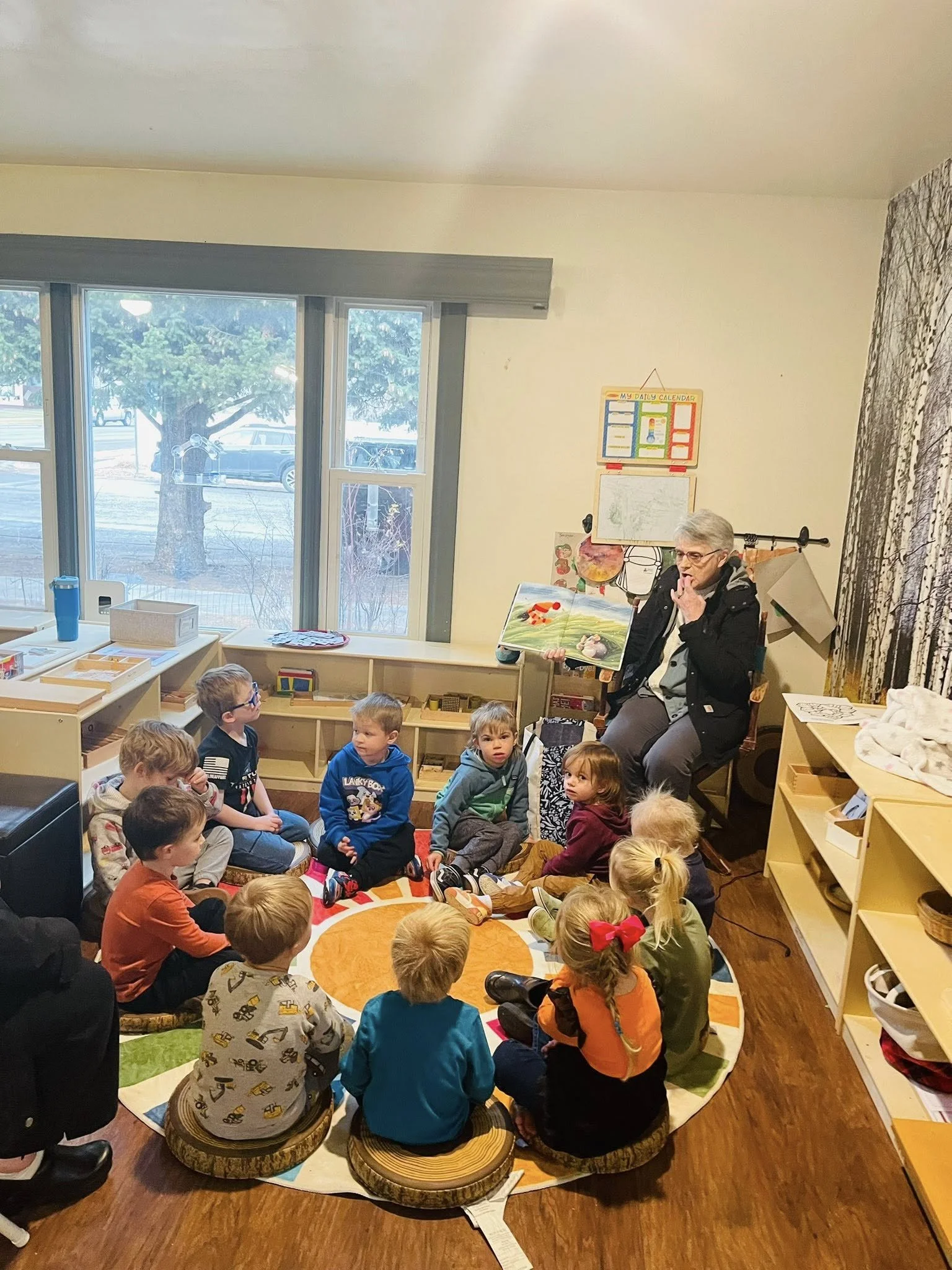 A group of young children sitting on cushions in a circle on a colorful rug, listening to a woman reading a picture book in a classroom with large windows, shelves filled with educational materials, and a mural of trees on the wall.