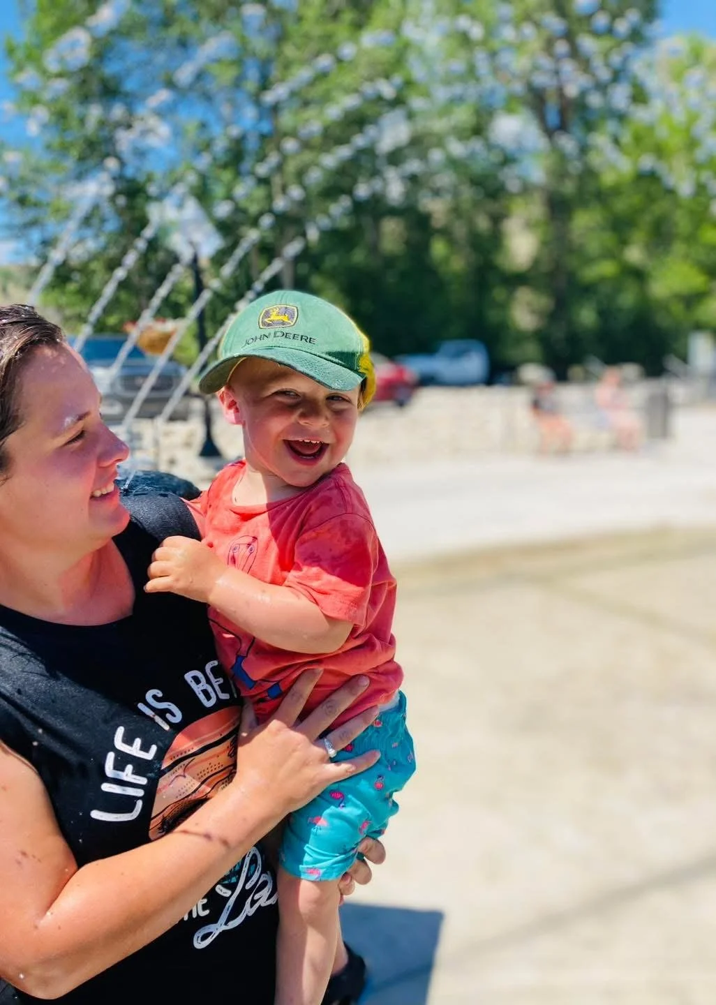 A smiling woman holding a happy young boy wearing a green John Deere cap outdoors on a sunny day.