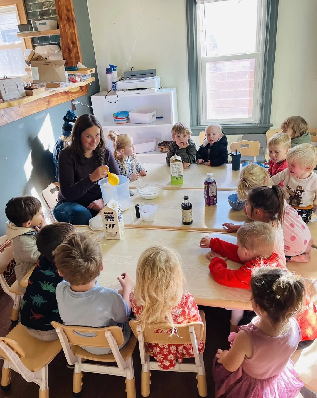 A woman leading a baking activity at a table surrounded by young children in a classroom or daycare setting.