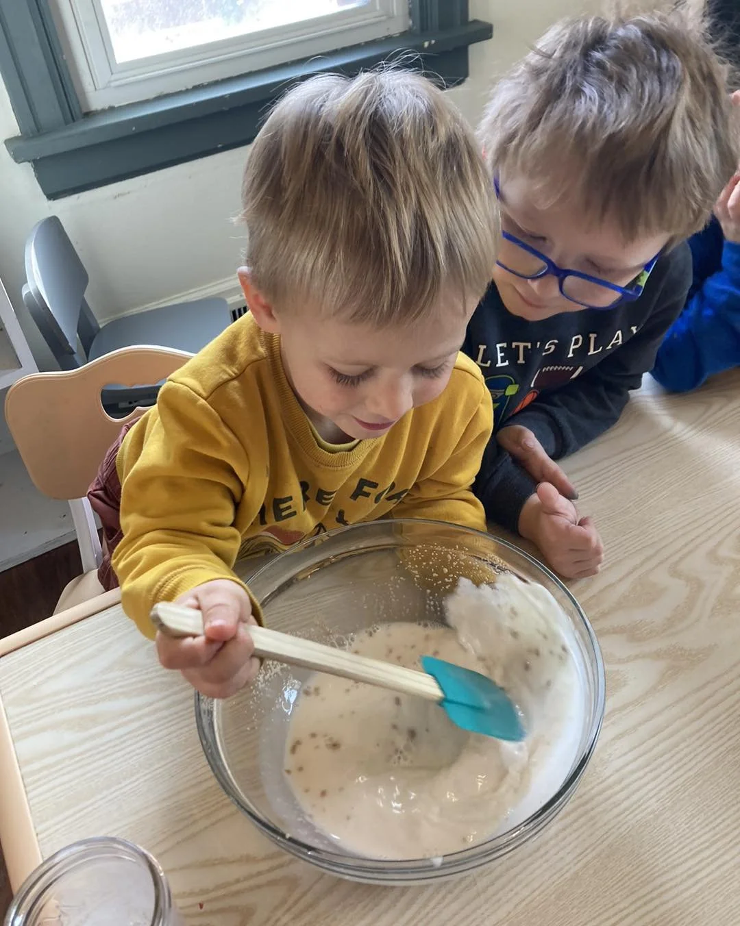 Two young boys with light hair and wearing glasses are leaning over a glass mixing bowl on a wooden table, with one boy stirring a mixture inside the bowl, while the other boy watches closely.