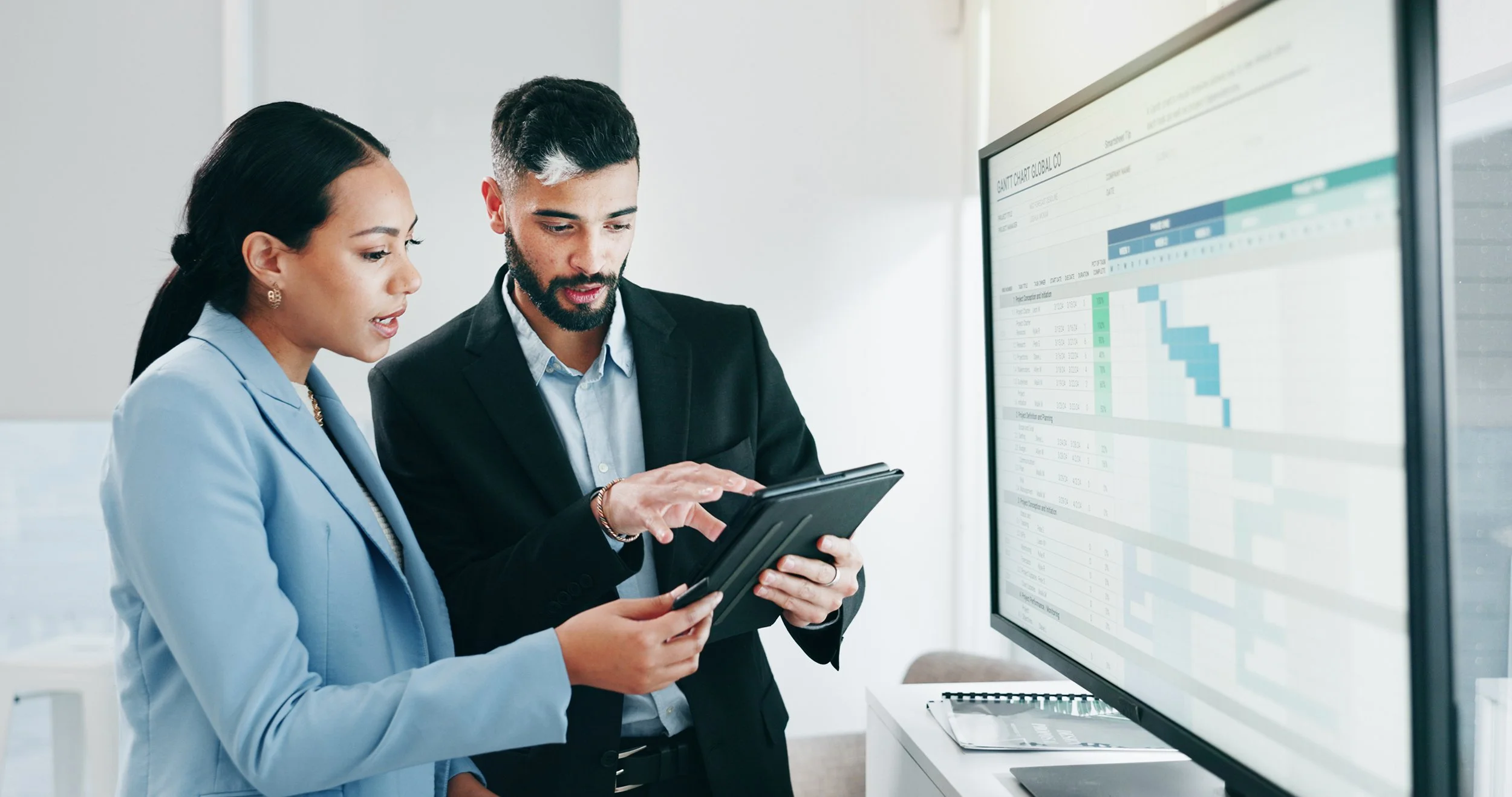 A woman and a man discussing information on a tablet with a large monitor in front of them with showing a GANTT chart.