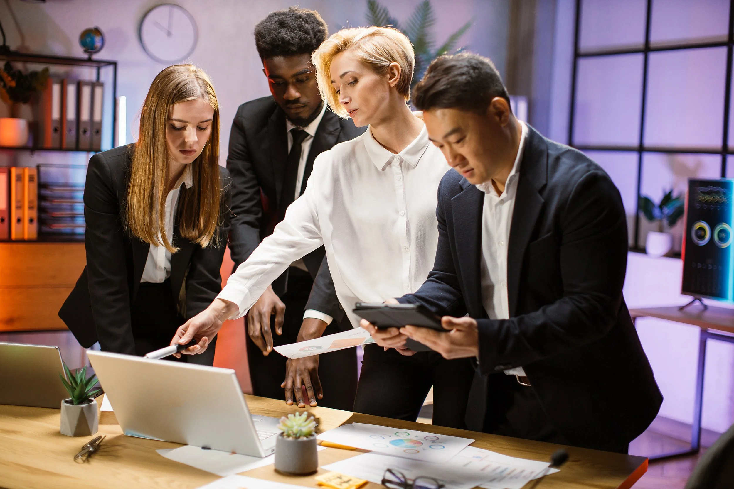 Business team of four discussing and analyzing with one women pointing at a laptop screen and holding a paper with information and one man holding an tablet.