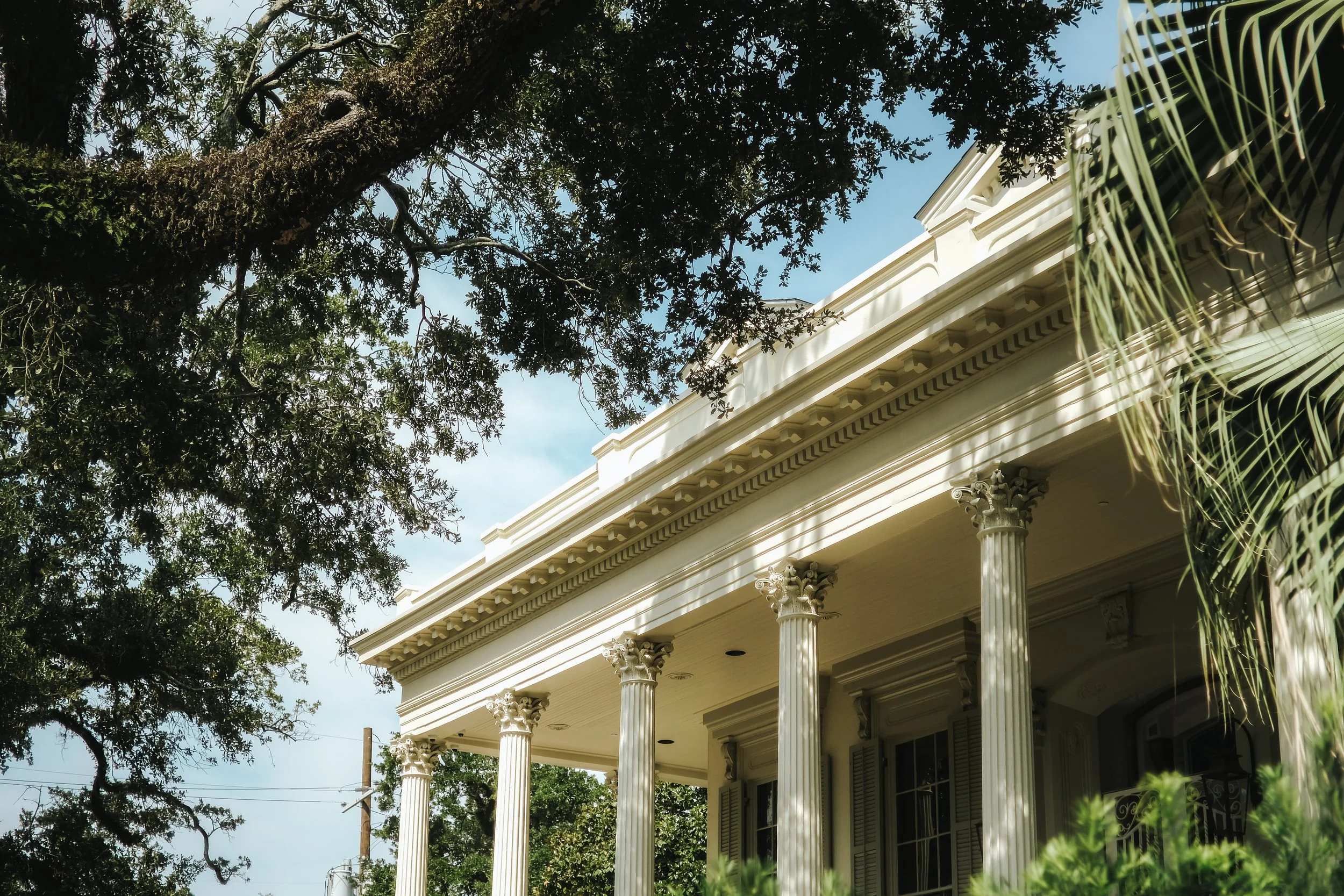 A white classical building with large columns and decorative frills, surrounded by green trees, under a partly cloudy sky.