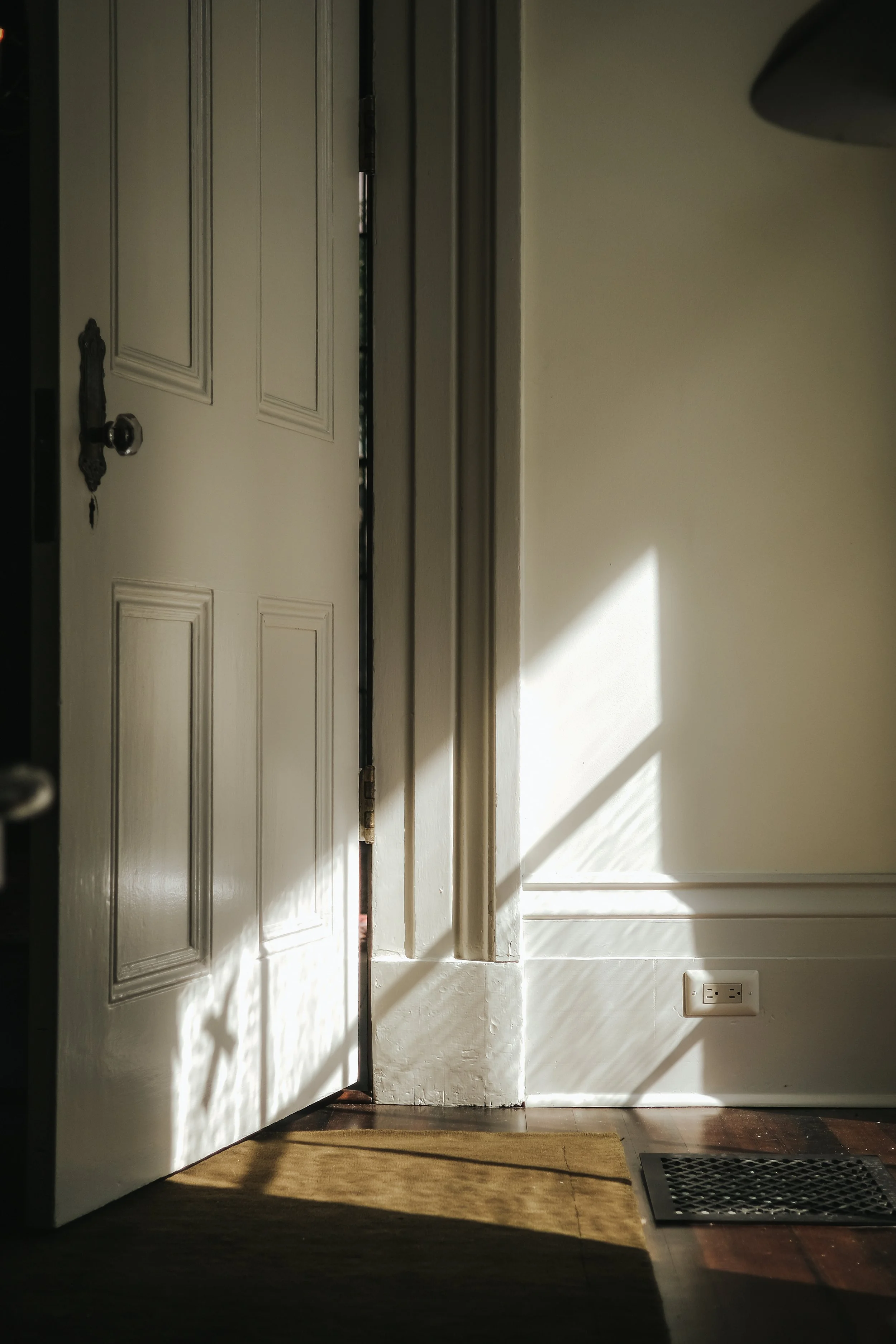 An open door with sunlight streaming in, revealing a white wall with a power outlet and a vent on the floor, with shadows cast by the sun.