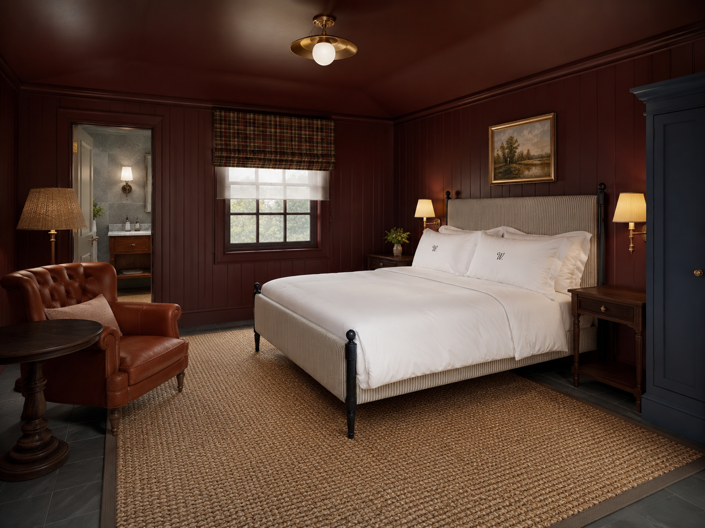 A cozy bedroom with dark wooden paneled walls, a white upholstered bed with monogrammed pillows, bedside lamps, a leather armchair, a window with plaid and sheer curtains, and a door leading to a bathroom.