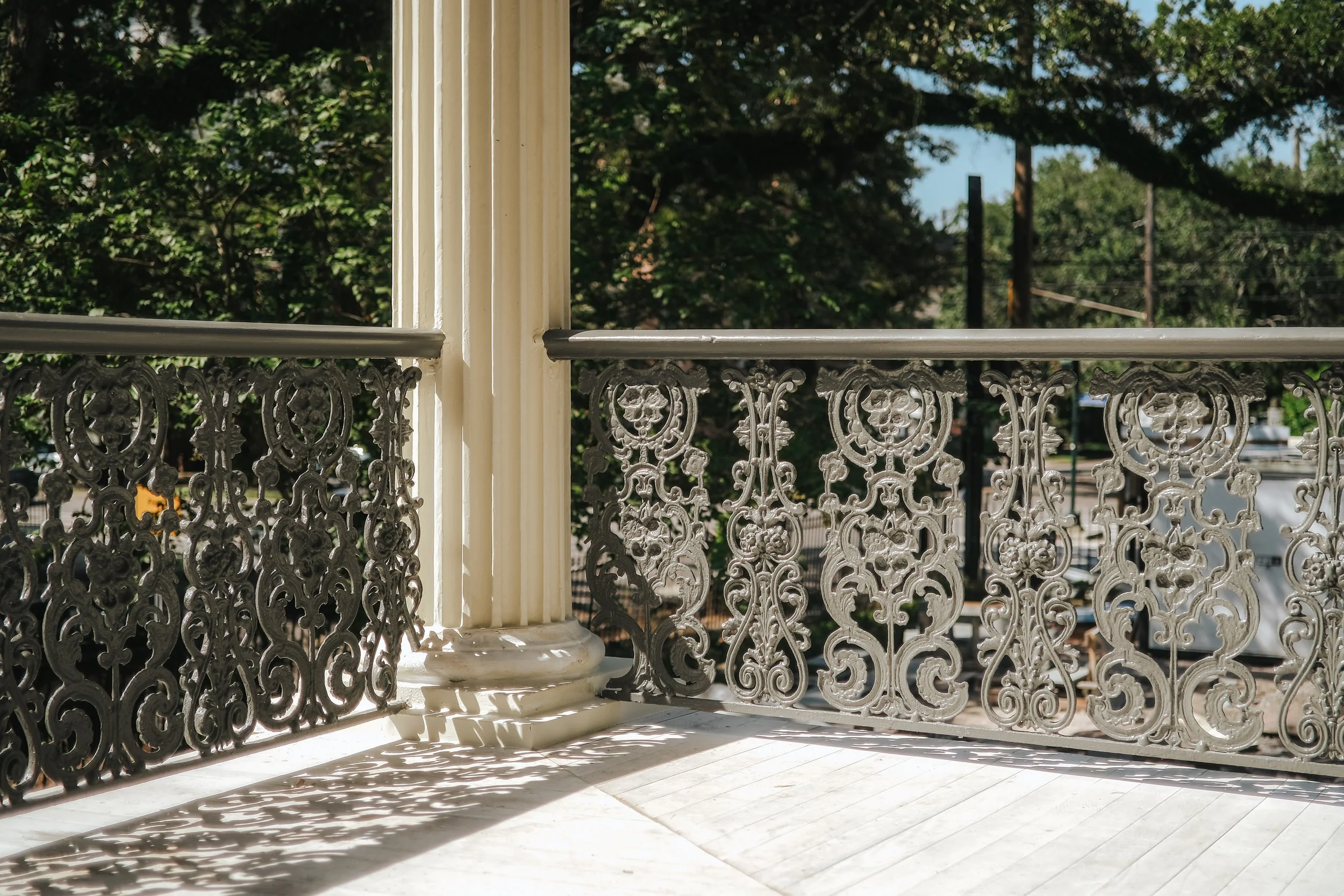 A decorative white metal railing with floral and swirl patterns, cast shadows on a white tiled porch, with a background of green trees and a clear blue sky.
