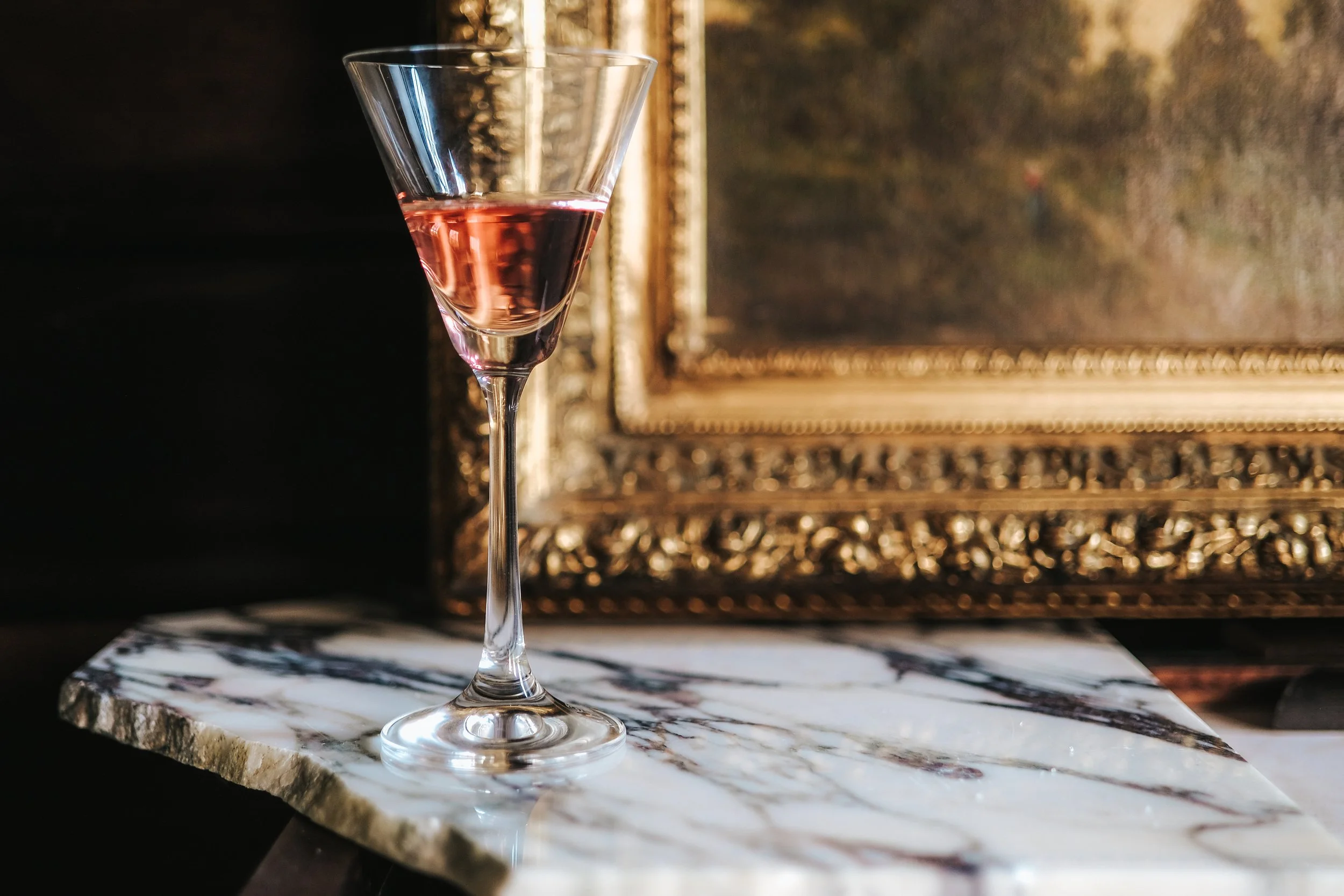 A martini glass with pink liquid on a marble surface, with a gold ornate framed painting in the background.