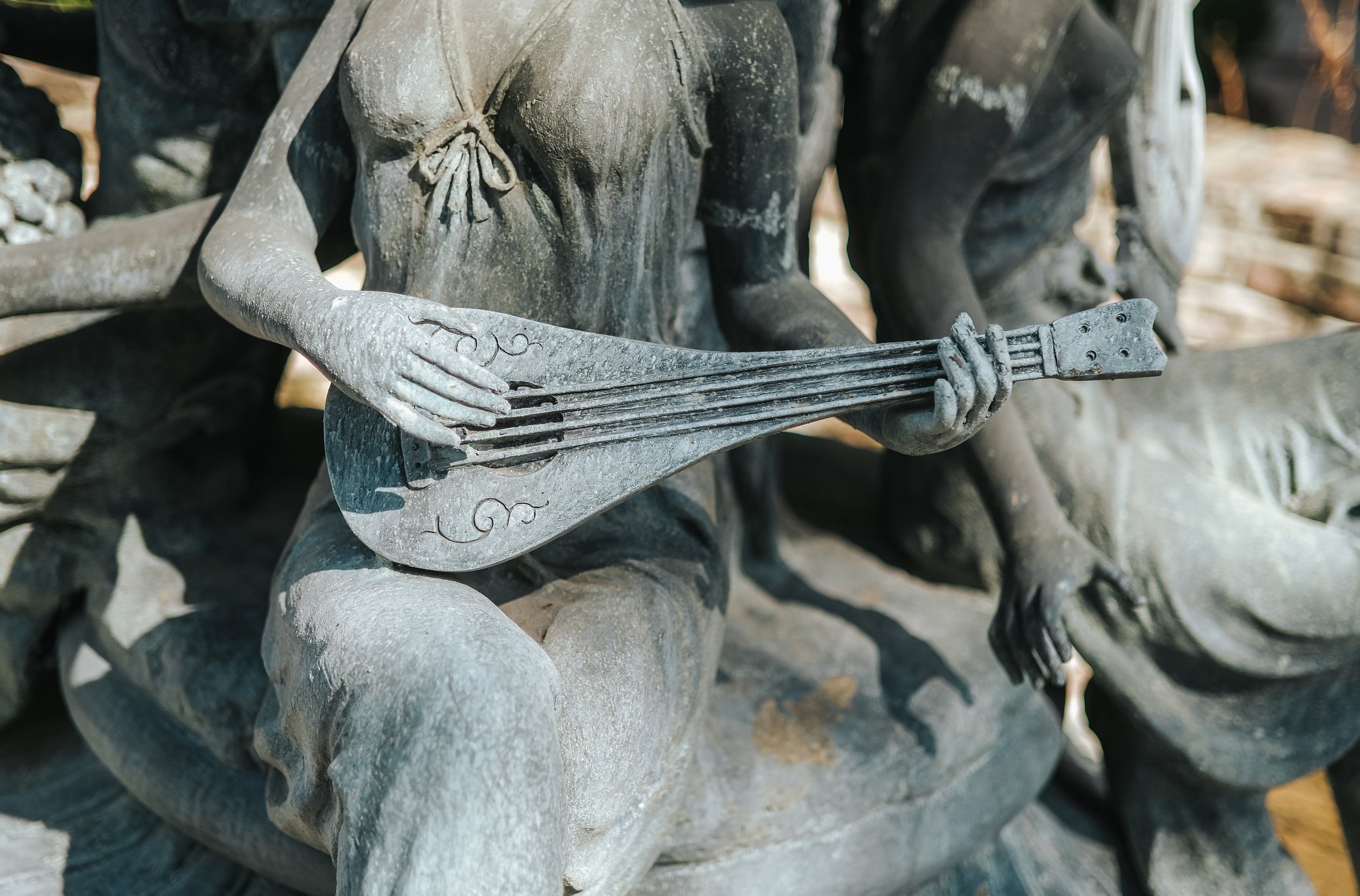 Close-up of a weathered metal sculpture of a person holding a guitar, showing detailed craftsmanship in the hands and torso, with a background of rocks.
