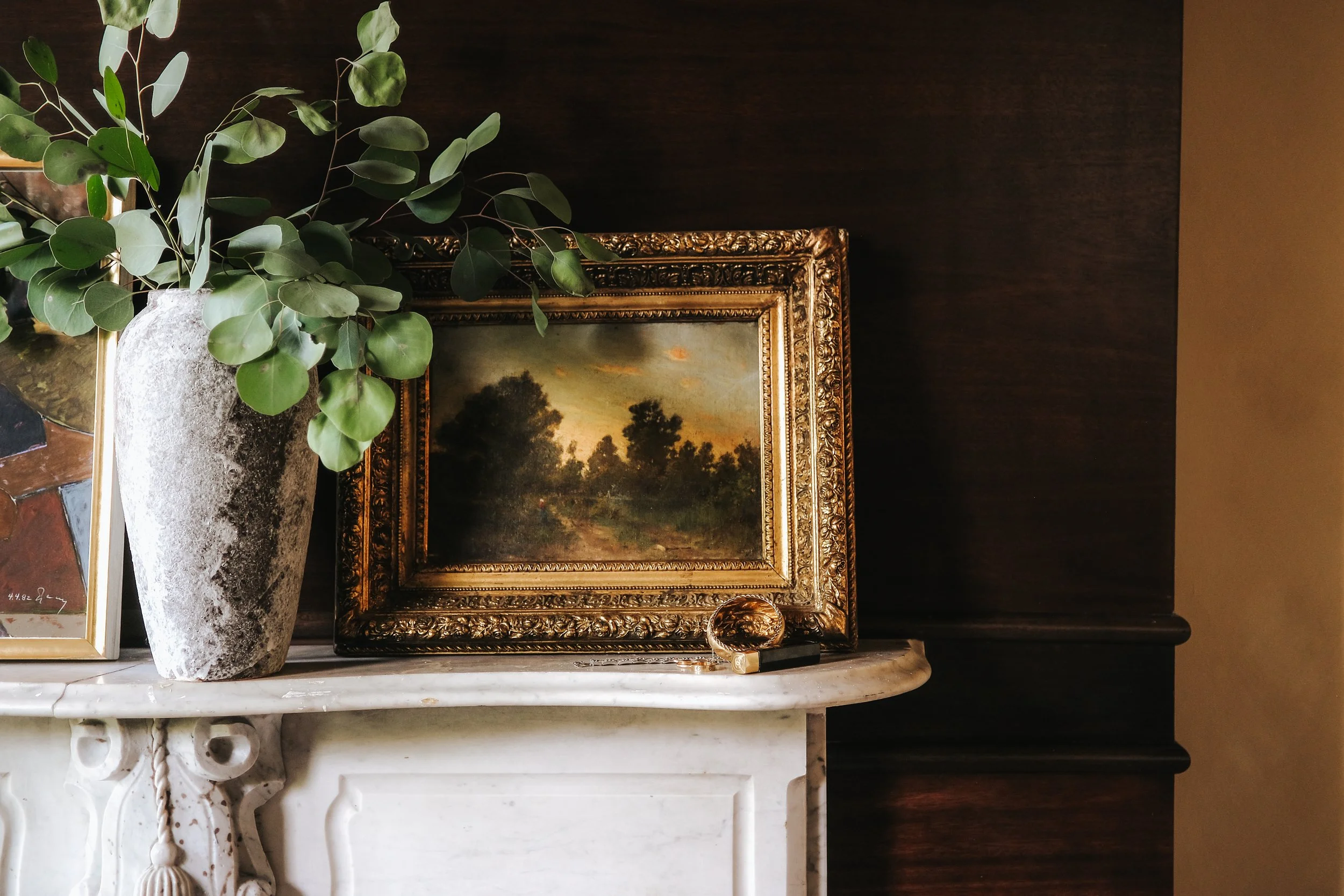 A tall potted plant with large green leaves positioned on a white marble mantelpiece. Behind the plant is a framed landscape painting with a gold ornate frame, depicting a sunset over trees. Part of a smaller picture frame is visible to the left of the plant.