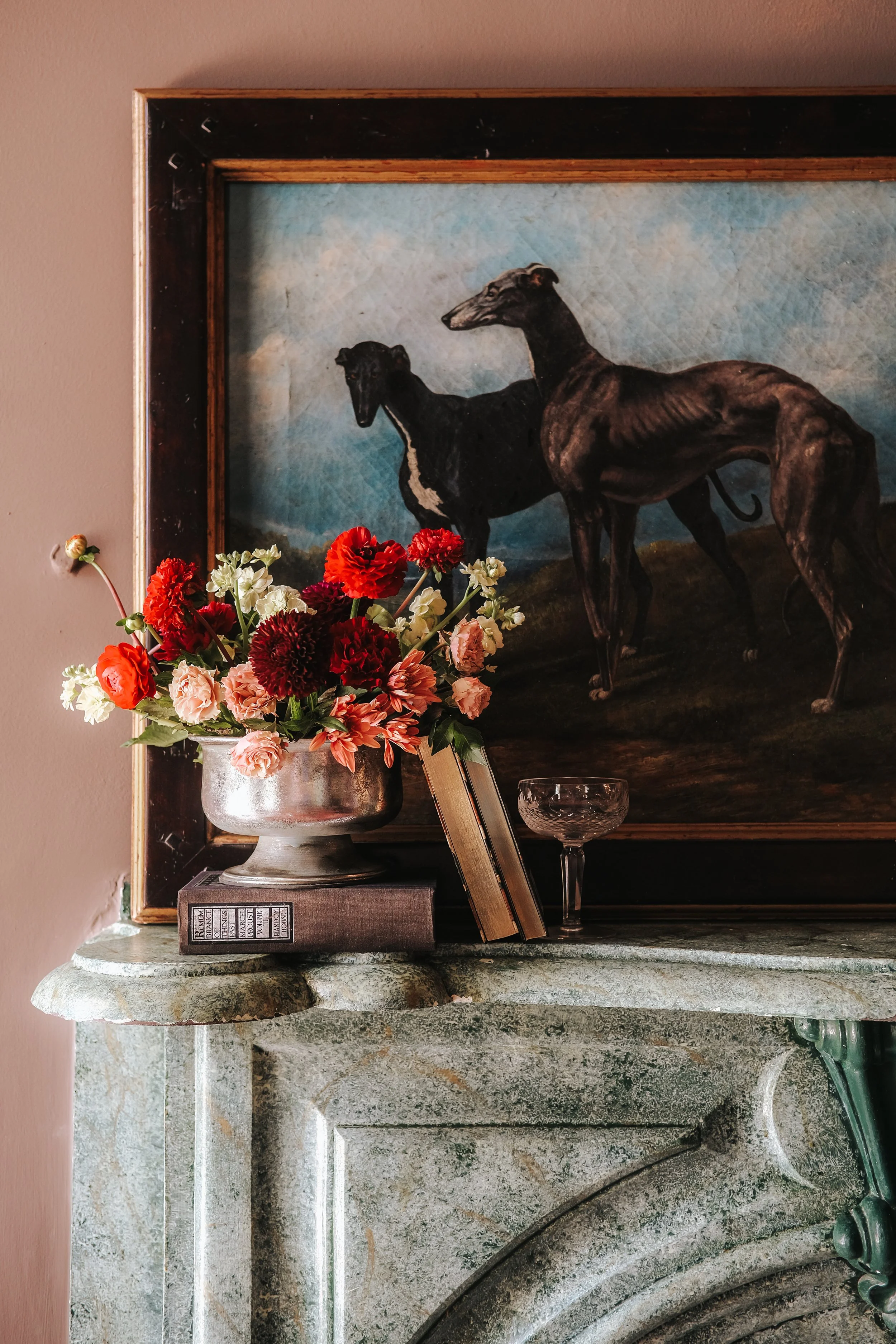 A floral arrangement of red, white, pink, and orange flowers in a silver vase placed on top of some books on a marble pedestal. Behind the flowers, there is a framed painting of two greyhound dogs against a sky with clouds.
