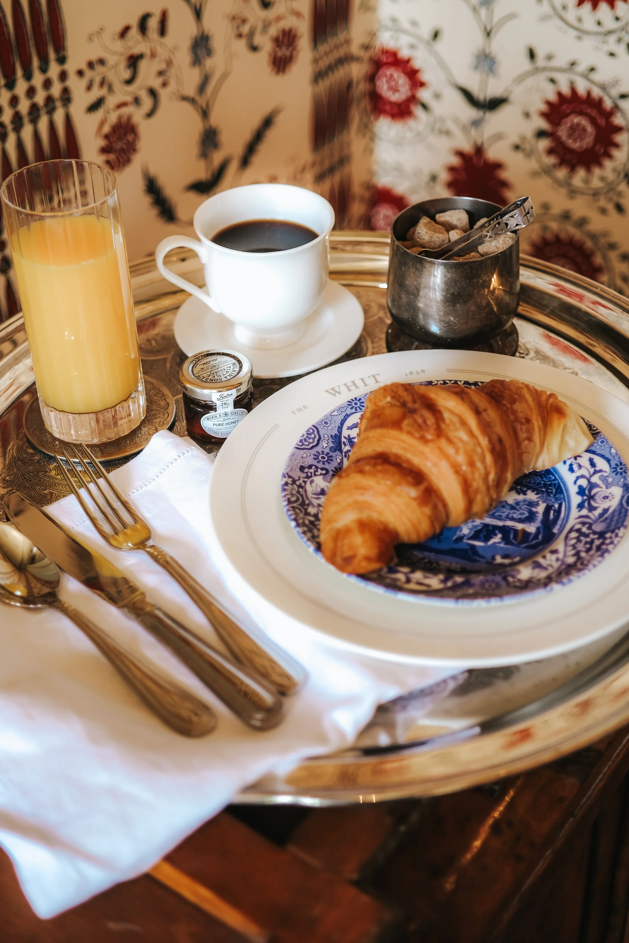 A breakfast tray with a croissant on a decorative plate, a cup of black coffee, a glass of orange juice, a small jar of honey, and a metal container of sugar cubes, set on a table with gold flatware.