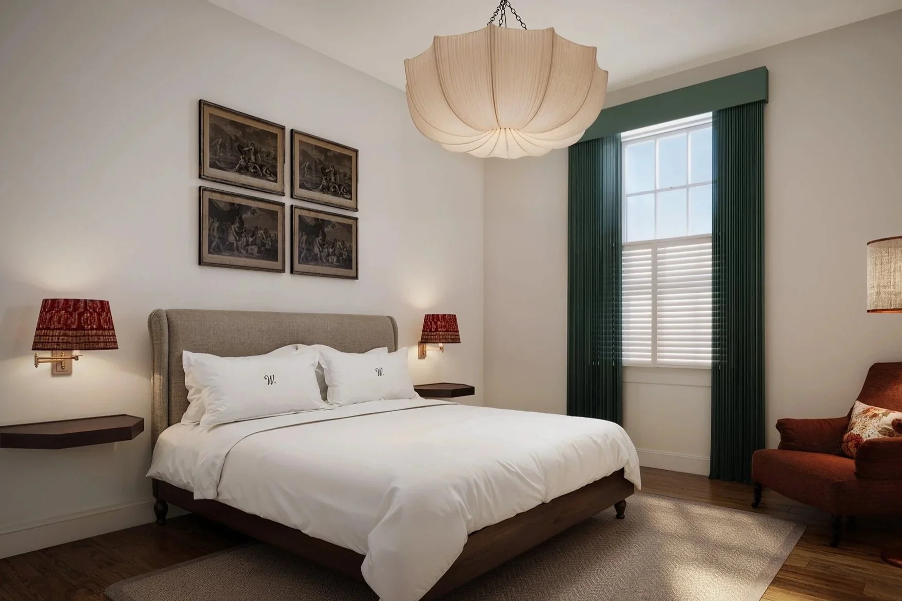 A bedroom with a beige upholstered bed, white bedding, and pillows with monograms, flanked by wall-mounted red lamps, and dark wood side tables. A window with white blinds and green curtains is on the right. Above the bed are four framed black-and-white photographs. There is a beige and brown rug on a wooden floor and a red armchair with a floral cushion in the corner.