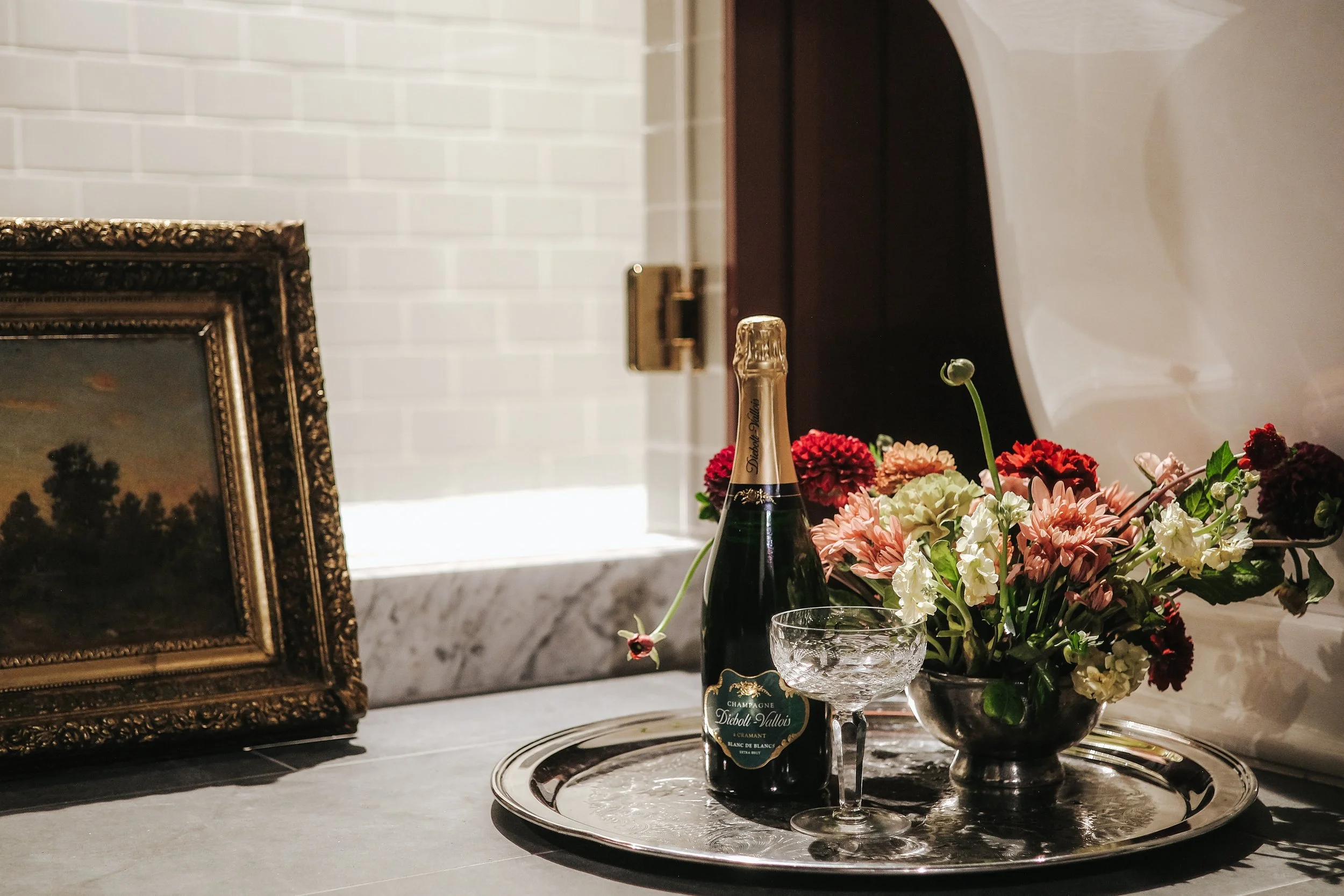 A still life of a champagne bottle, a floral arrangement, an ornate picture frame, and a glass on a tray on a countertop, with a white tiled wall in the background.