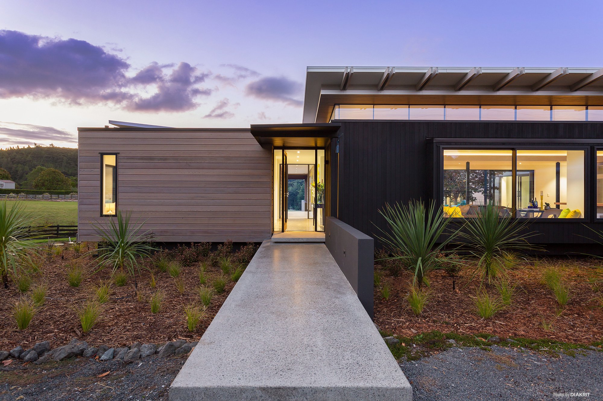 Modern house with large windows, black and gray exterior, lit interior, surrounded by landscaped yard with plants, under a partly cloudy sky during sunset.