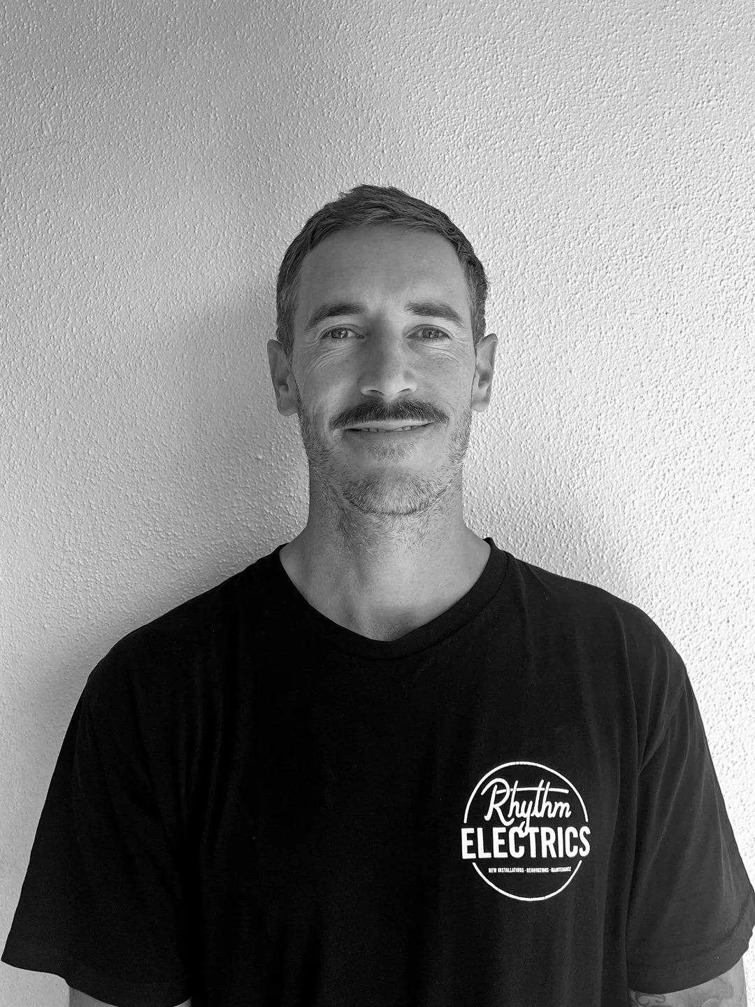 Black and white photo of a man with short hair and a beard, wearing a black T-shirt with the logo 'Rhythm ELECTRICS' on the chest, standing in front of a plain wall.