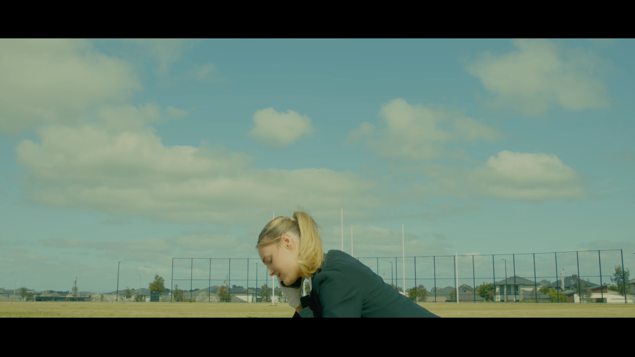 A woman in a black blazer sitting on the ground, speaking on a cellphone, in an outdoor green field with a fence and houses in the background, under partly cloudy sky.